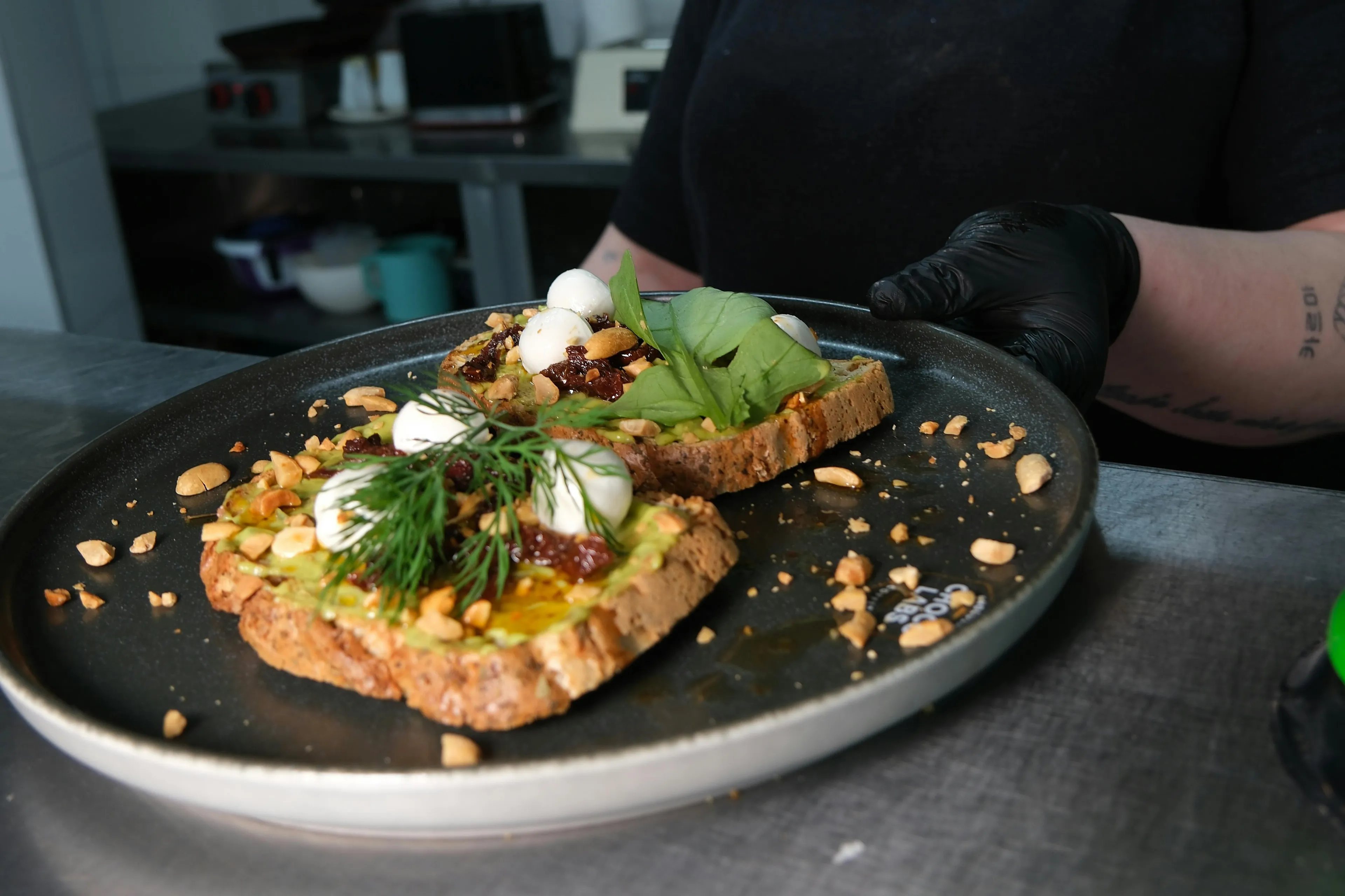 A chef wearing black gloves presents a plate with two slices of gourmet toast topped with avocado, mozzarella, and nuts. A comfort food option.