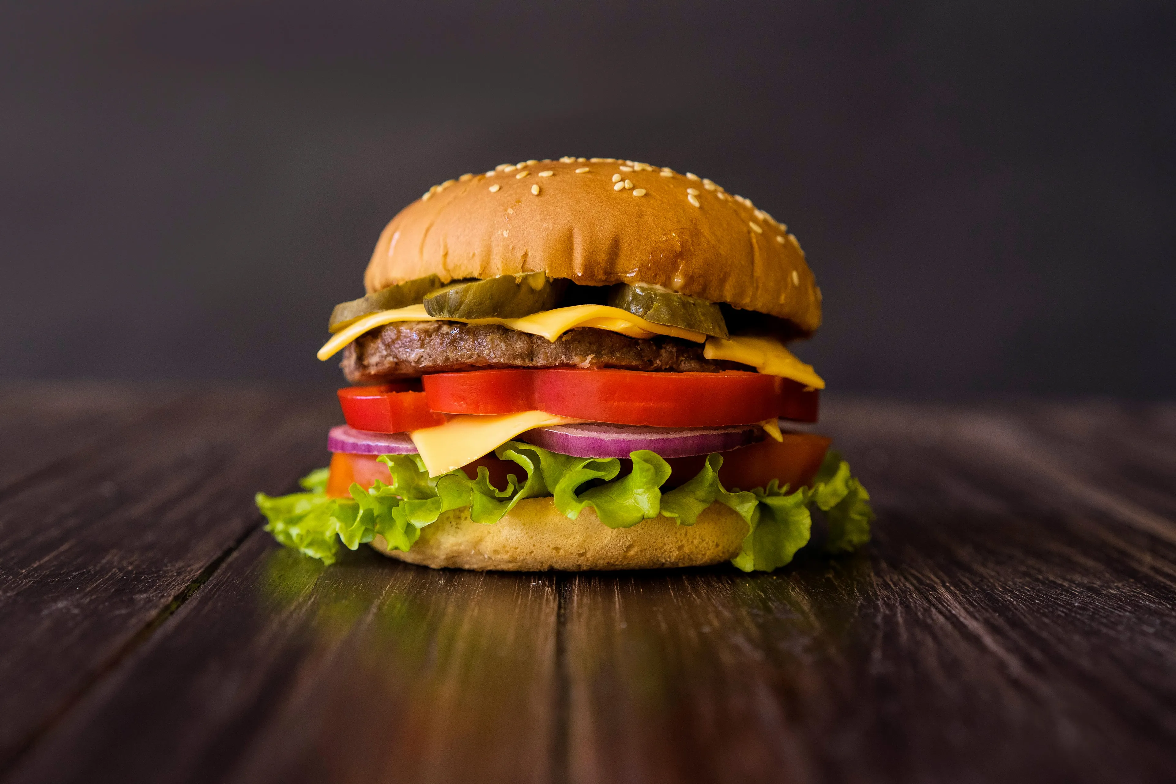 A delicious stacked Burger with cheese, pickles, tomatoes, onion, and lettuce on a dark wooden surface. Comfort food.