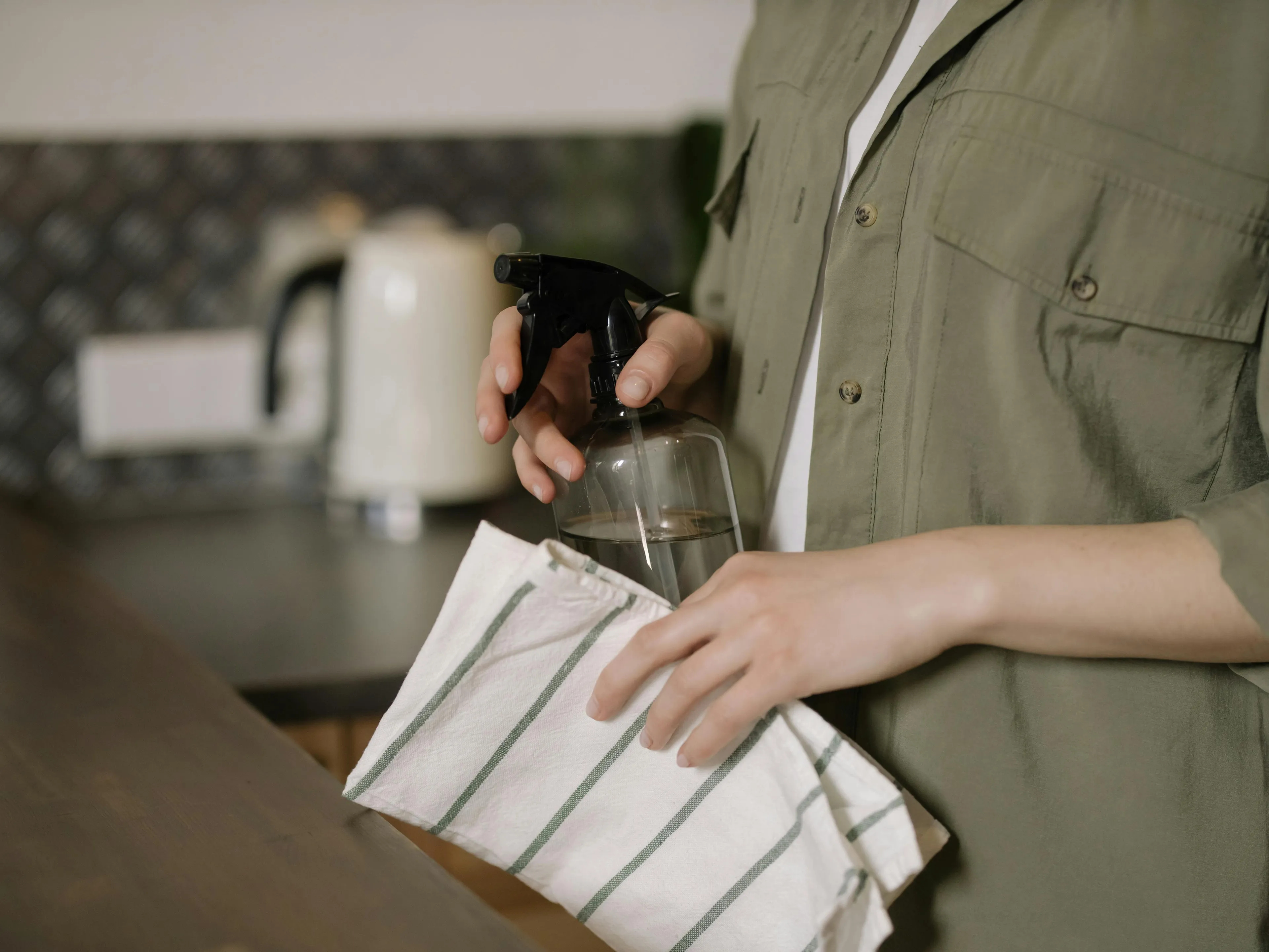 Hands holding a spray bottle and striped cloth, ready to deep clean the kitchen as a rainy-day activity.