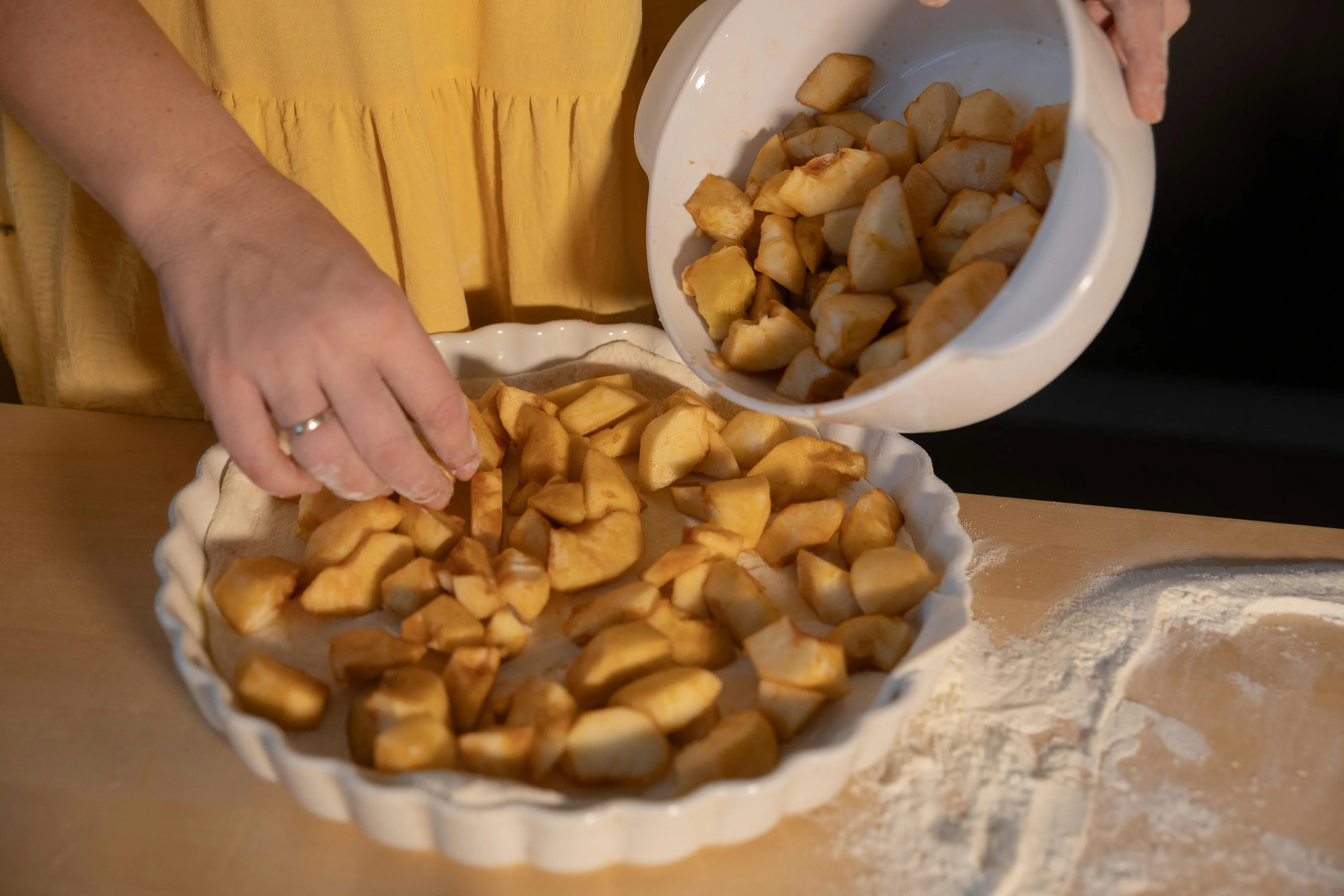 A person in a yellow shirt preparing to bake an apple pie, adding chopped apples to a pie crust in a white dish.