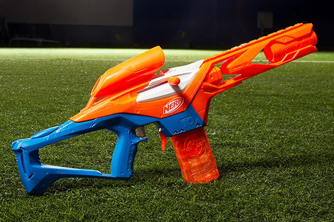 A vibrant orange and blue Nerf Blaster lies on green artificial turf, ready for action.