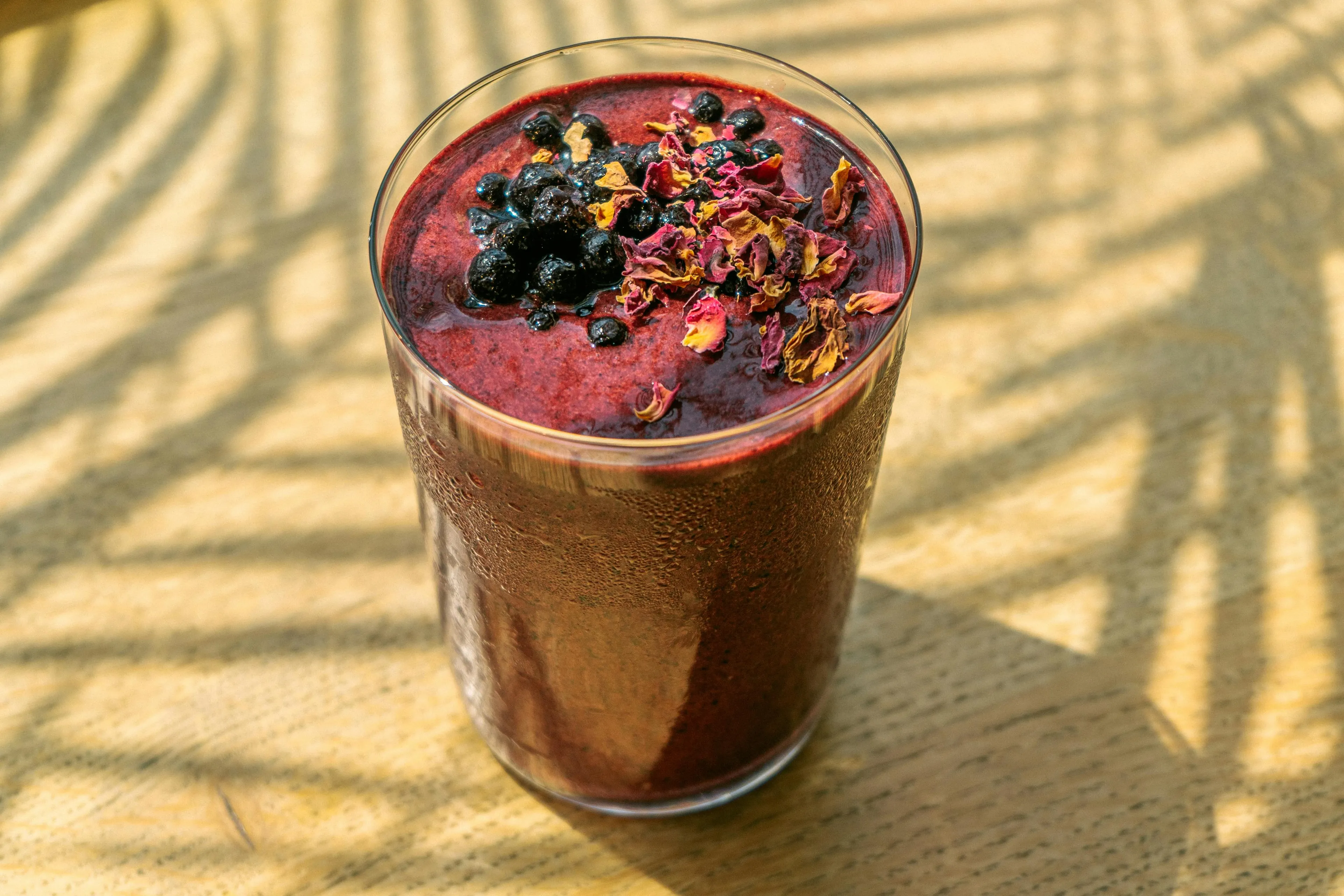 A chilled protein smoothie, topped with blueberries and rose petals, sits on a wooden table with dappled sunlight.