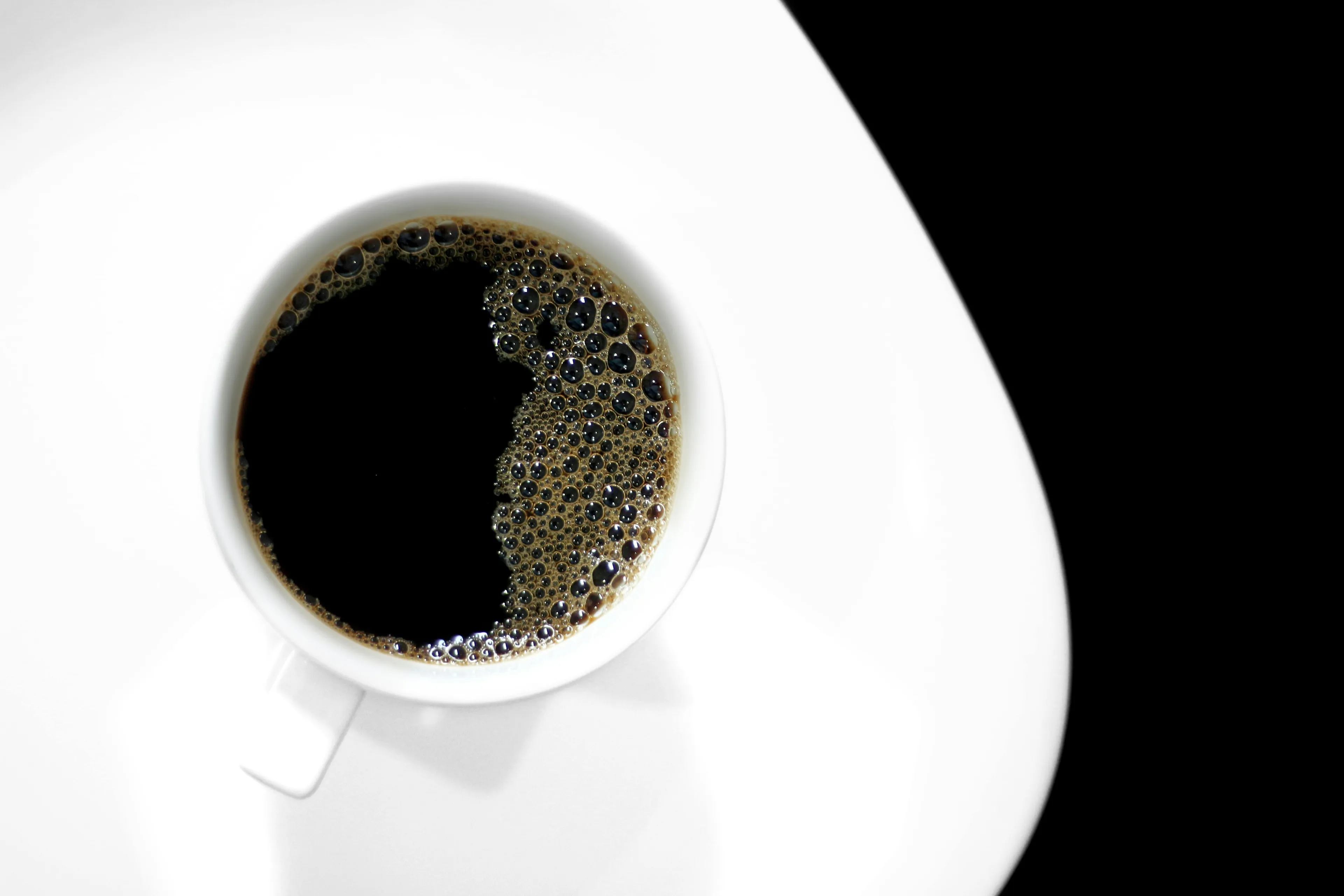 A top-down view of black coffee in a white cup and saucer. Dark coffee, with a bubbly surface, against a black background.