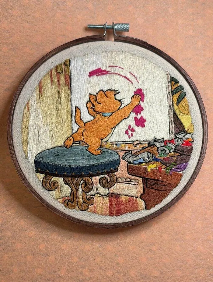 An embroidery project showing an orange kitten painting pink paw prints on a canvas
