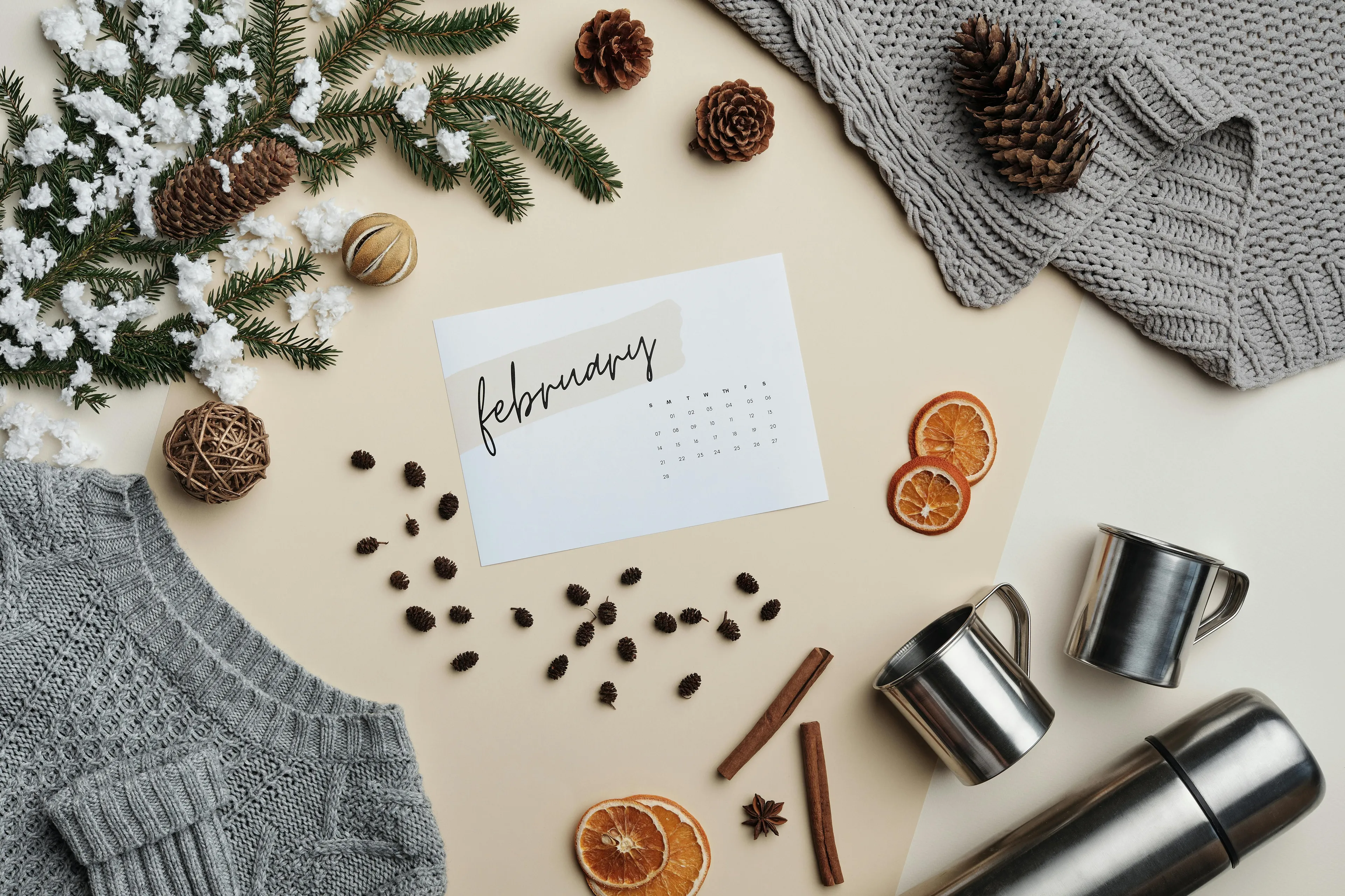 Overhead shot of February calendar, pinecones, snow-dusted pine branches, cozy grey knitwear, and travel mugs. Trivia night.