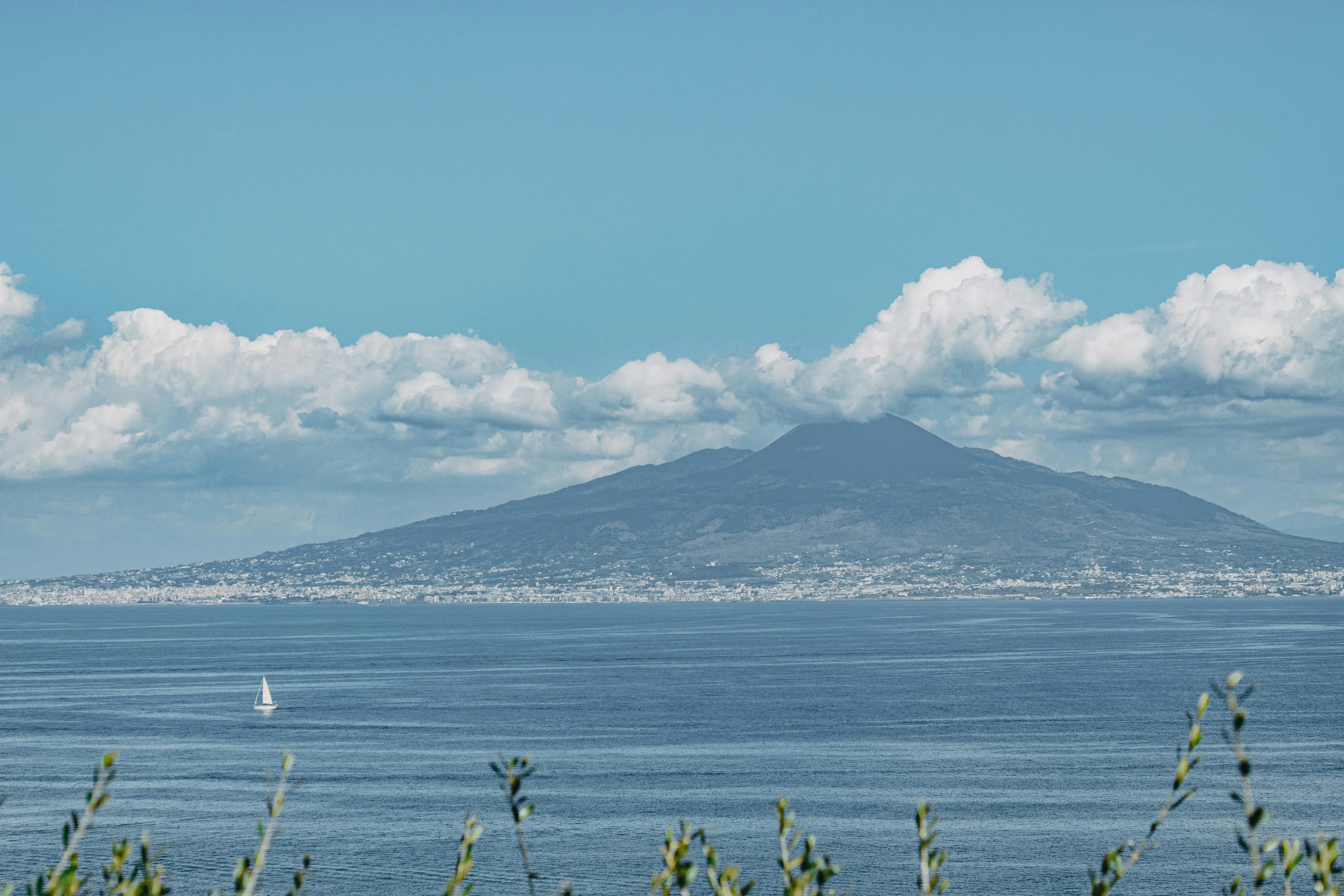 A scenic view of the Bay of Naples with Mount Vesuvius in the background, a sailboat on the water. Win trivia night.