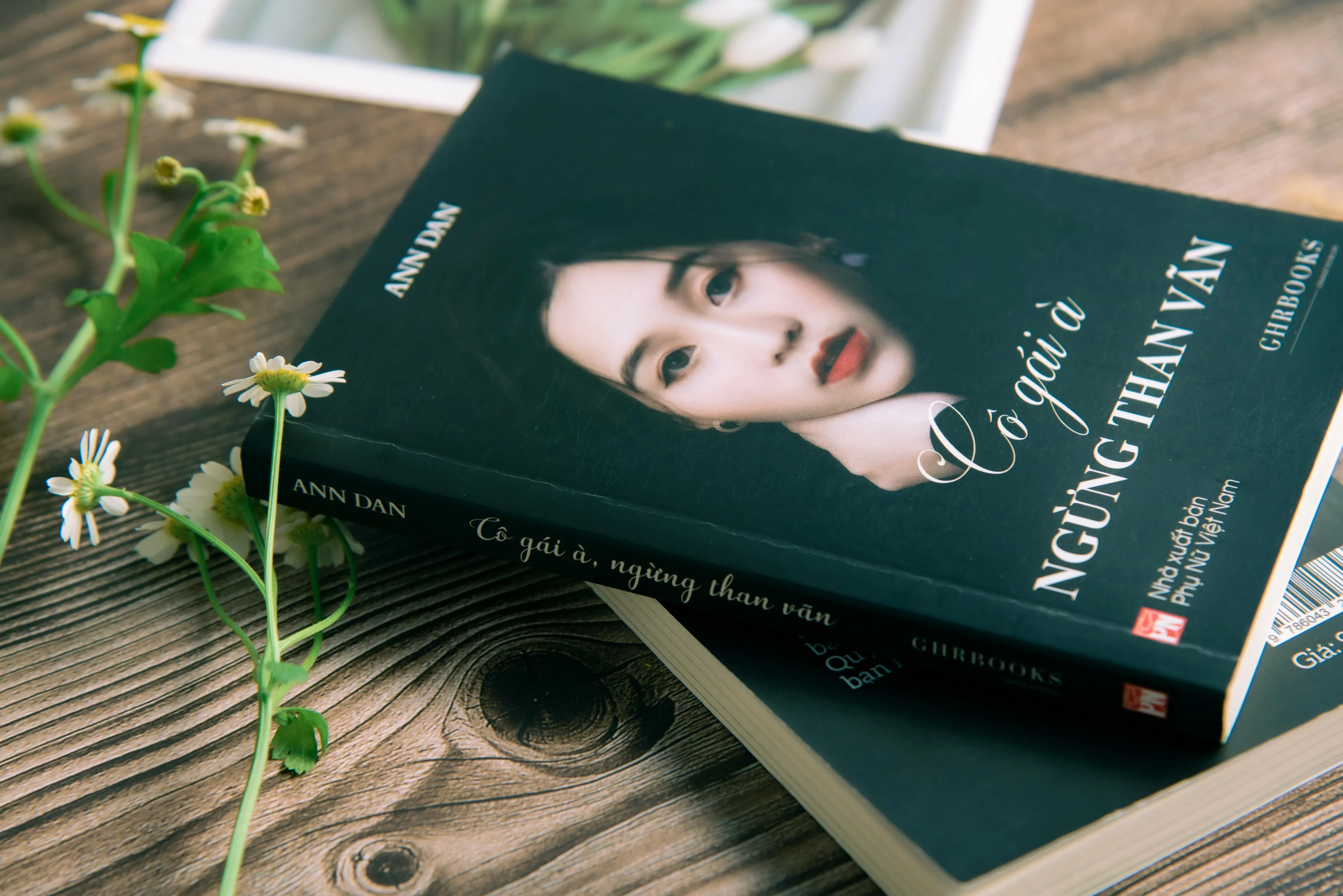 A book with a woman's face on the cover, lying on a wooden surface beside delicate white flowers. A perfect housewarming gift.