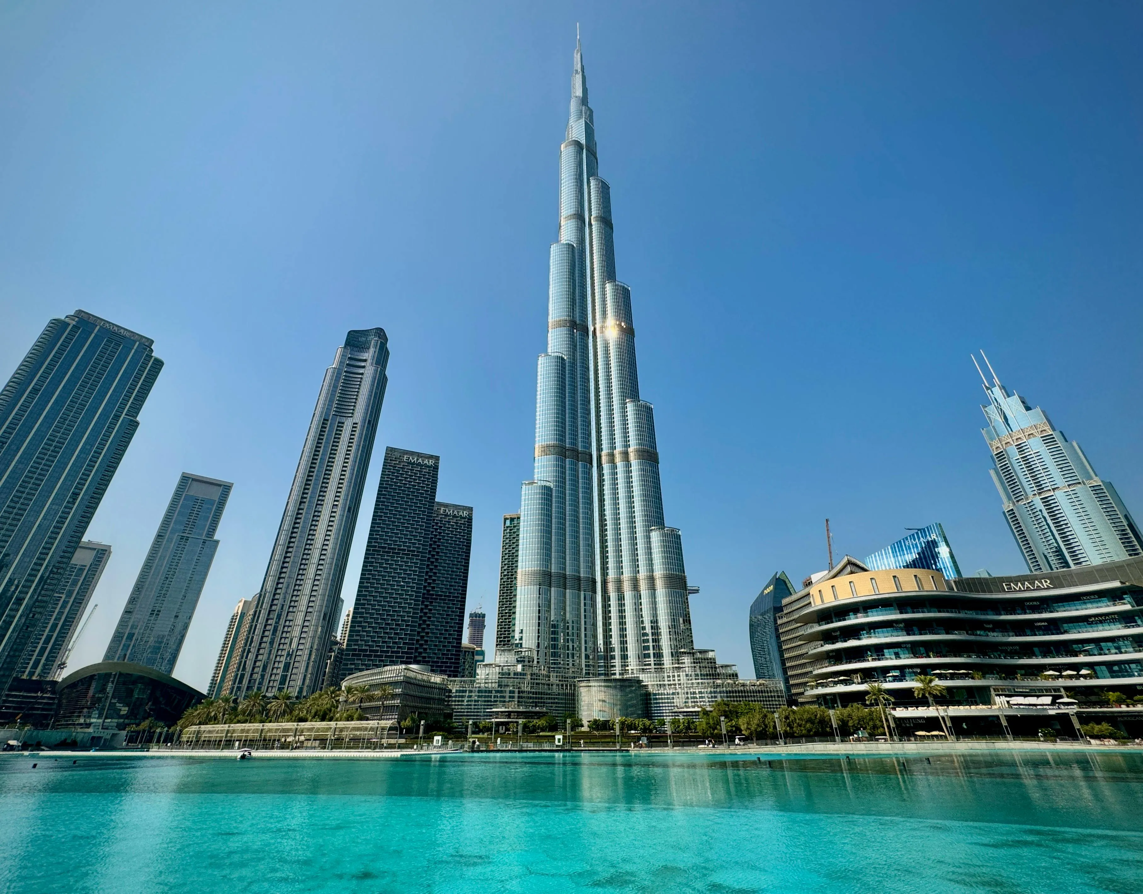 Burj Khalifa, majestic against a bright blue sky, rises above surrounding skyscrapers and a vibrant turquoise water feature.