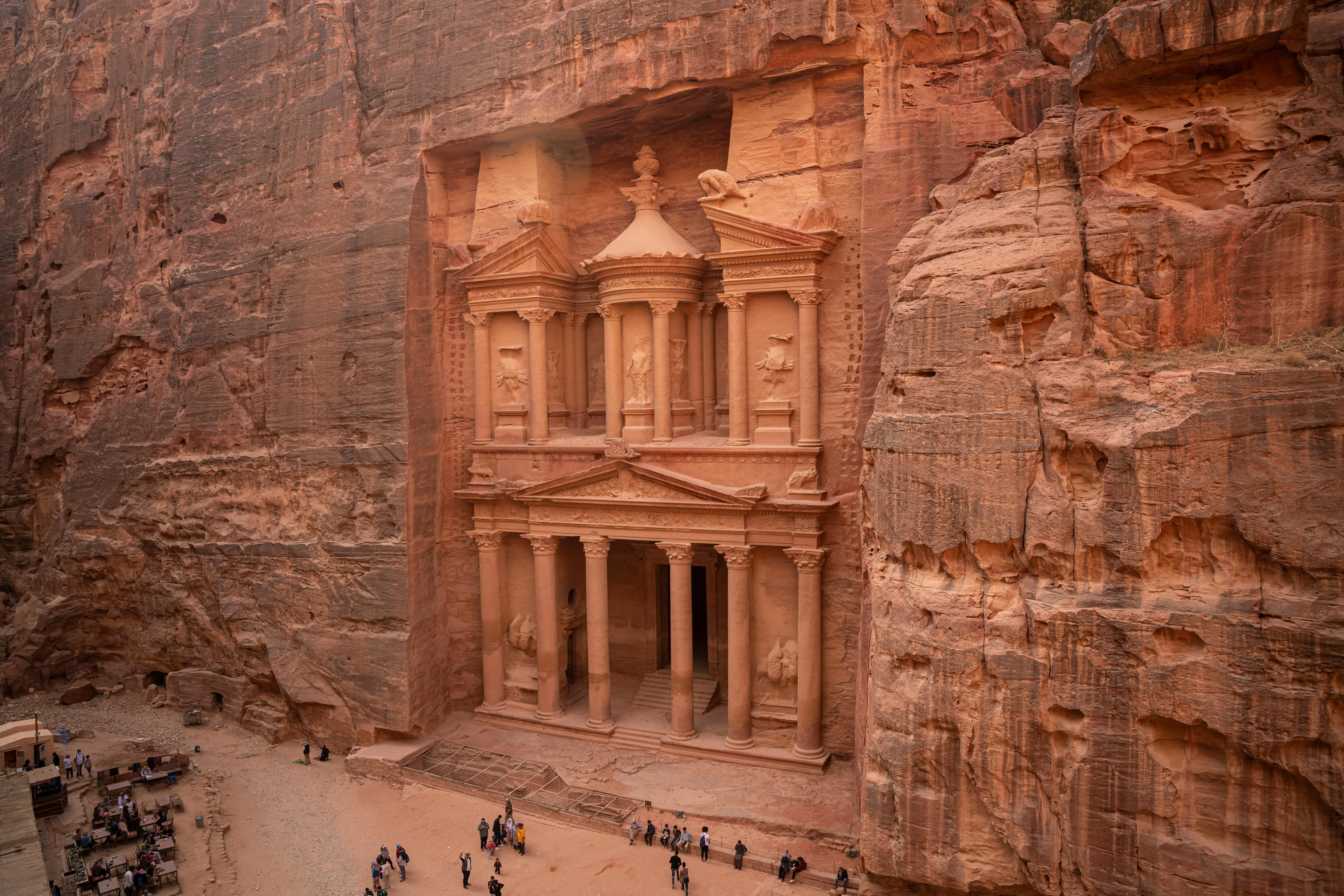 Panoramic view of the Treasury at Petra, an ancient rock-cut temple carved into a reddish cliff face, with tourists.