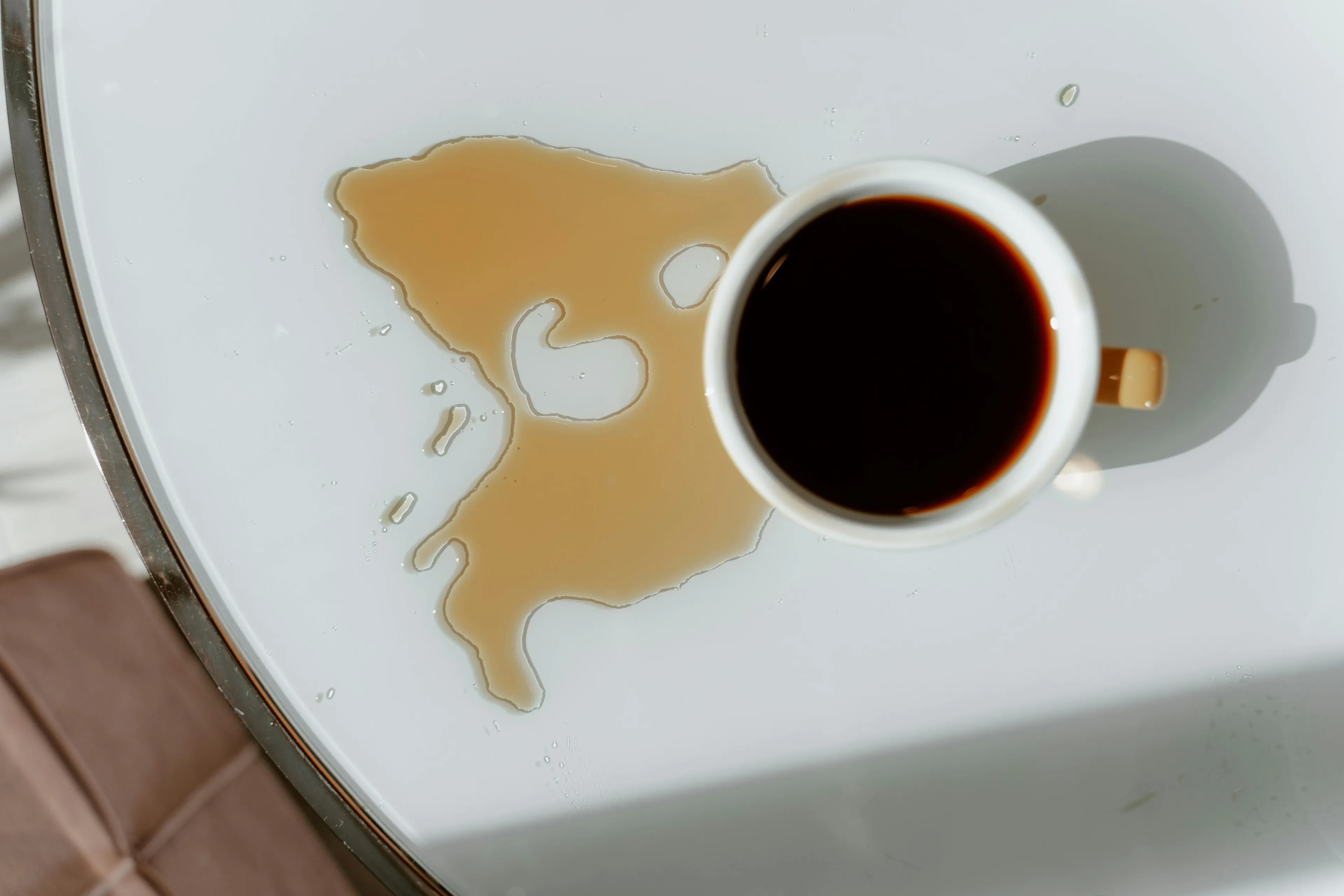 Overhead shot of spilled coffee on a white table, next to a full coffee cup. Someone spills coffee, how do you apologize?