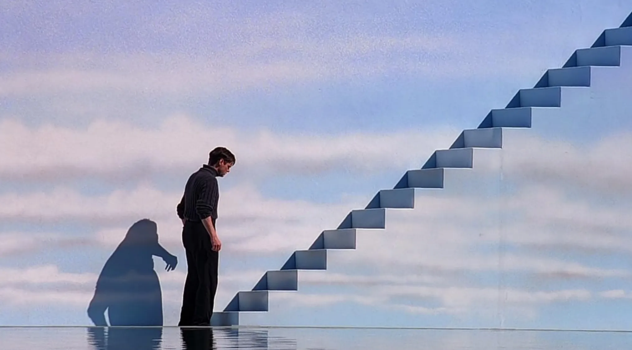 A man standing before a long staircase ascending into a cloudy sky, reflected in water. Movie quiz scene.