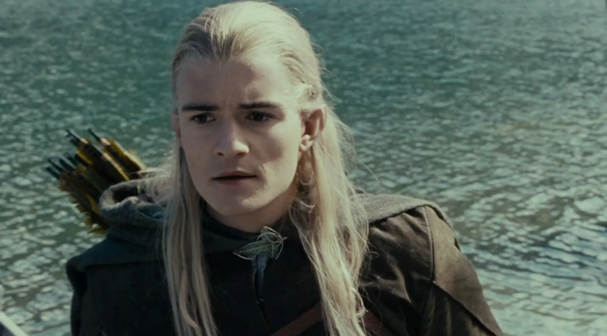 A close-up of the Lord of the Rings character, Legolas, with long blonde hair, looking serious by water. Movie quiz character.