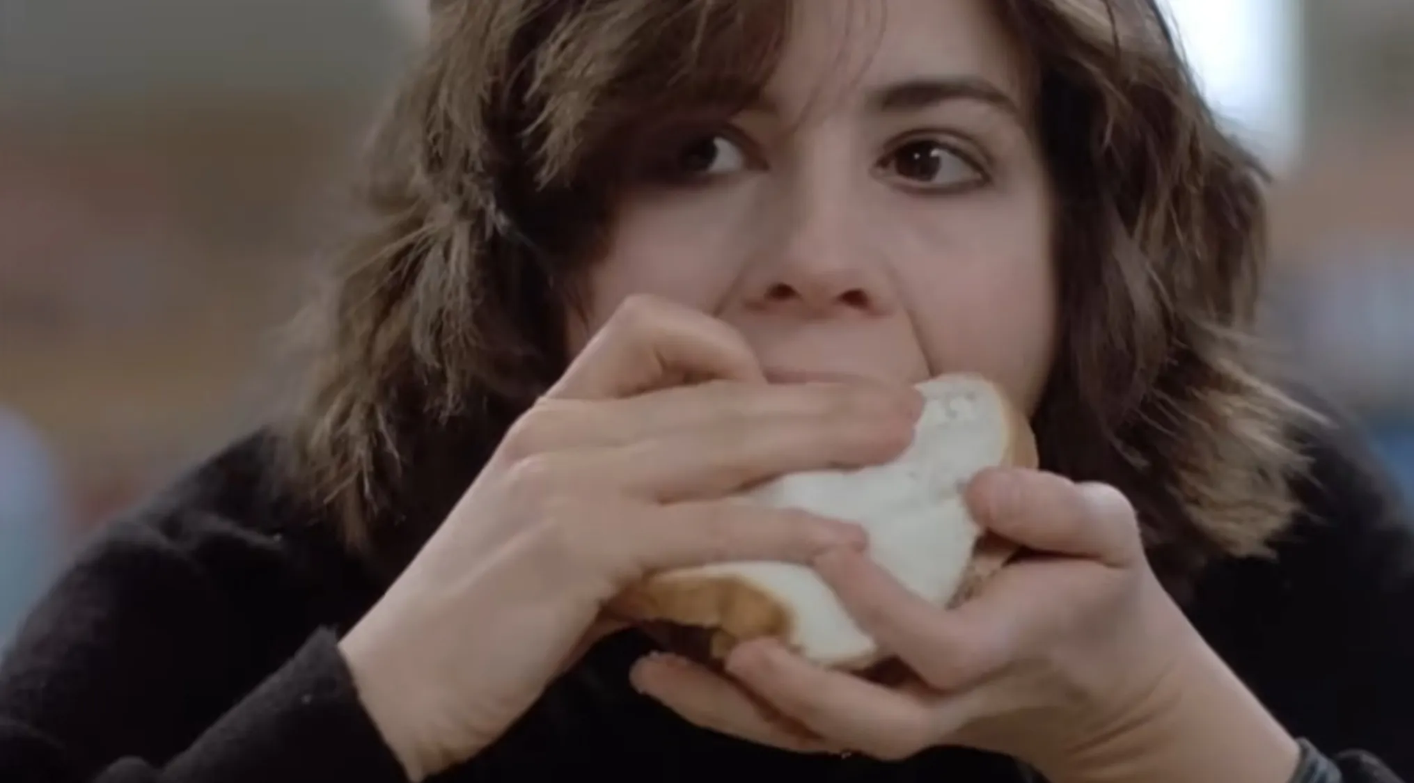 A young woman with brown hair intently eating a sandwich, looking upward. A movie quiz scene.