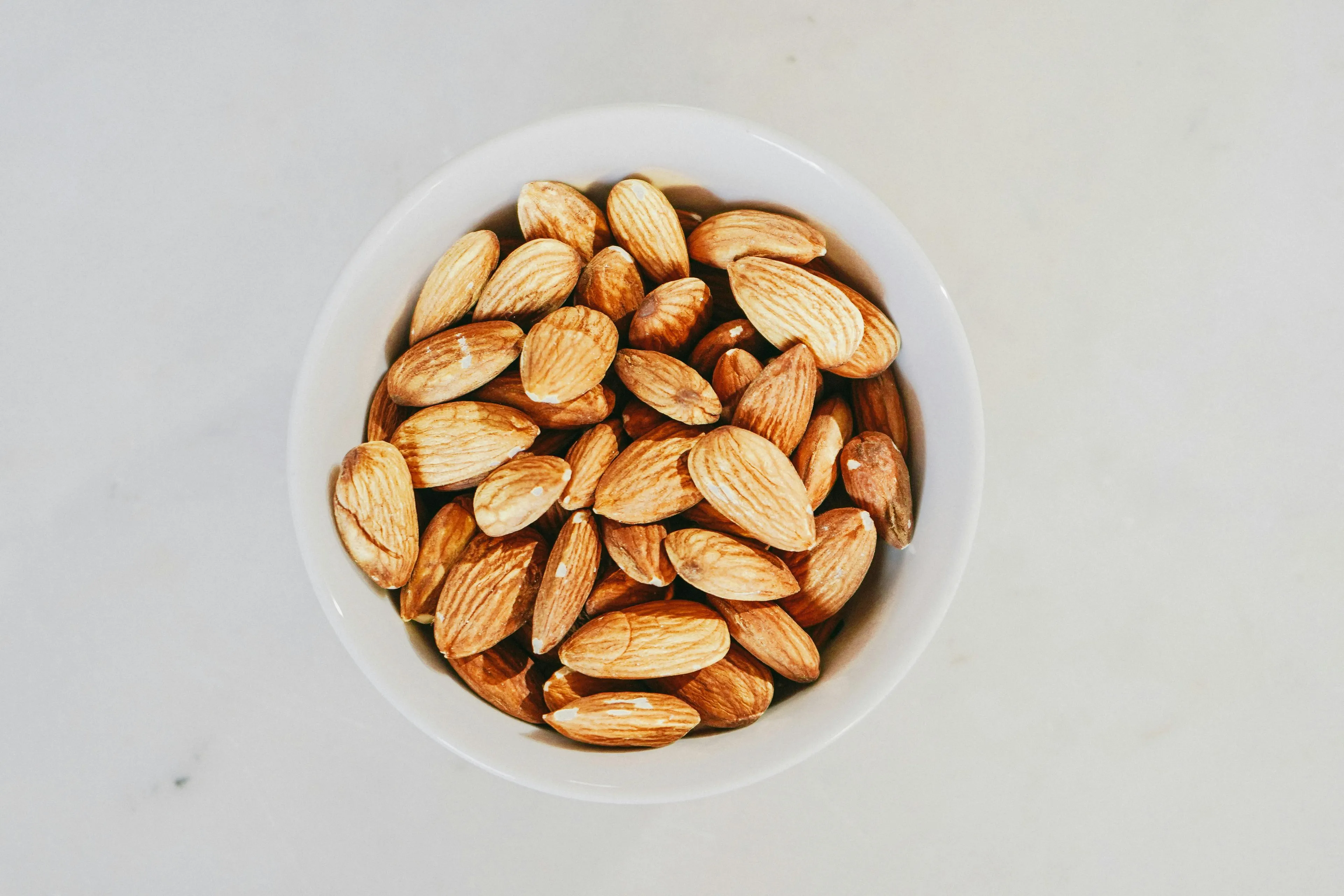 A white bowl filled with numerous almonds, a perfect comfort snack, on a white background.