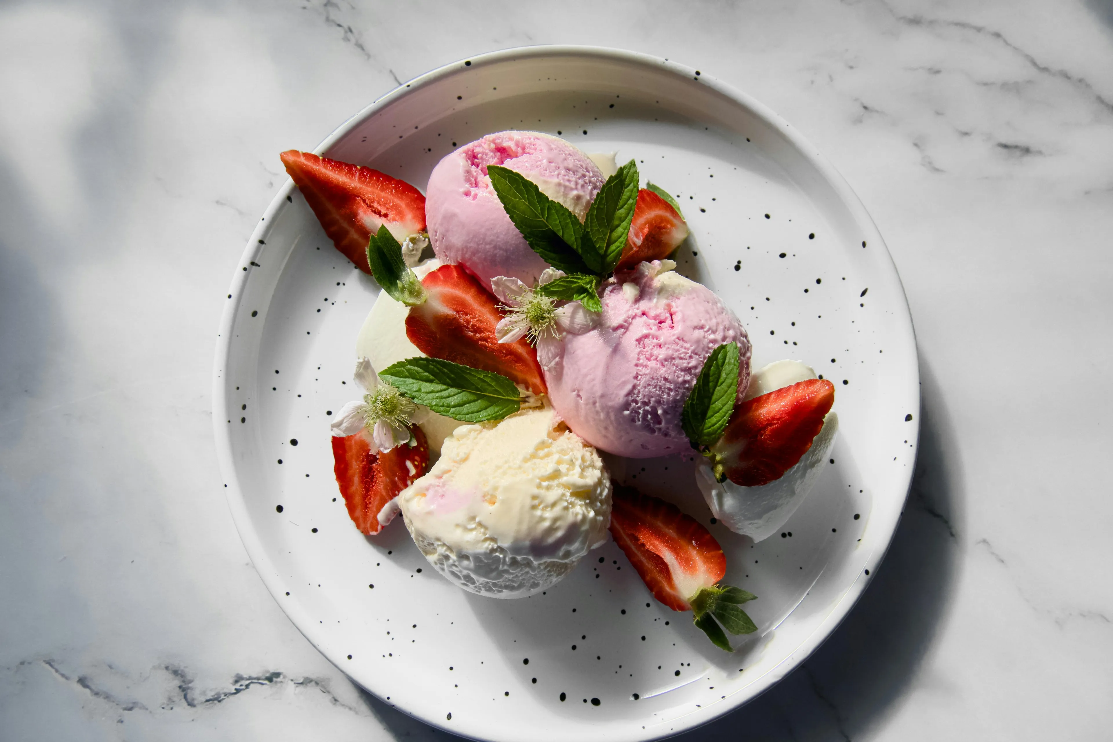 A gourmet serving of strawberry and vanilla ice cream, garnished with fresh mint leaves and sliced strawberries, on a white plate.