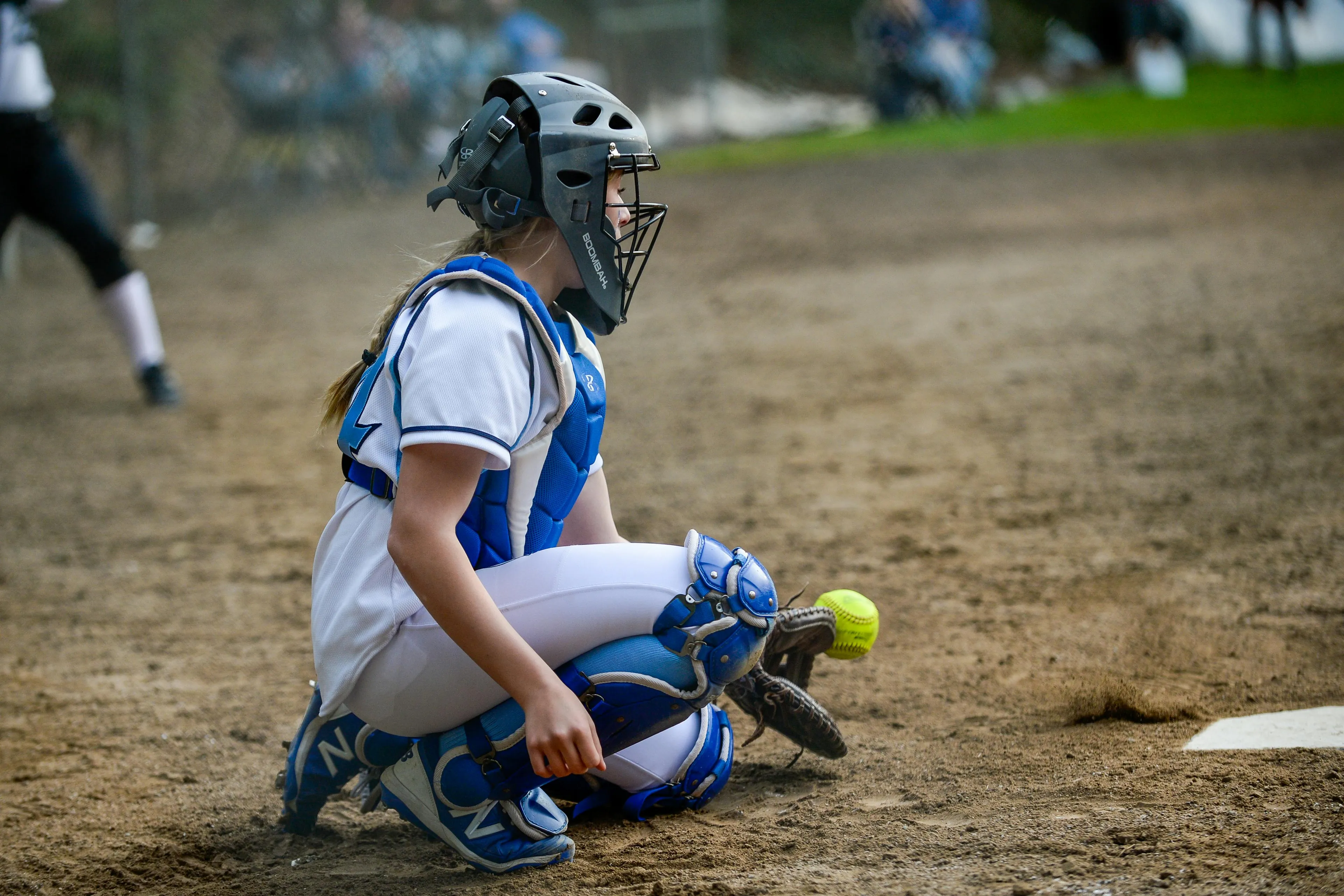 A young softball catcher in blue and white gear, kneeling in the dirt, catching a yellow softball. Reaction to rejection.