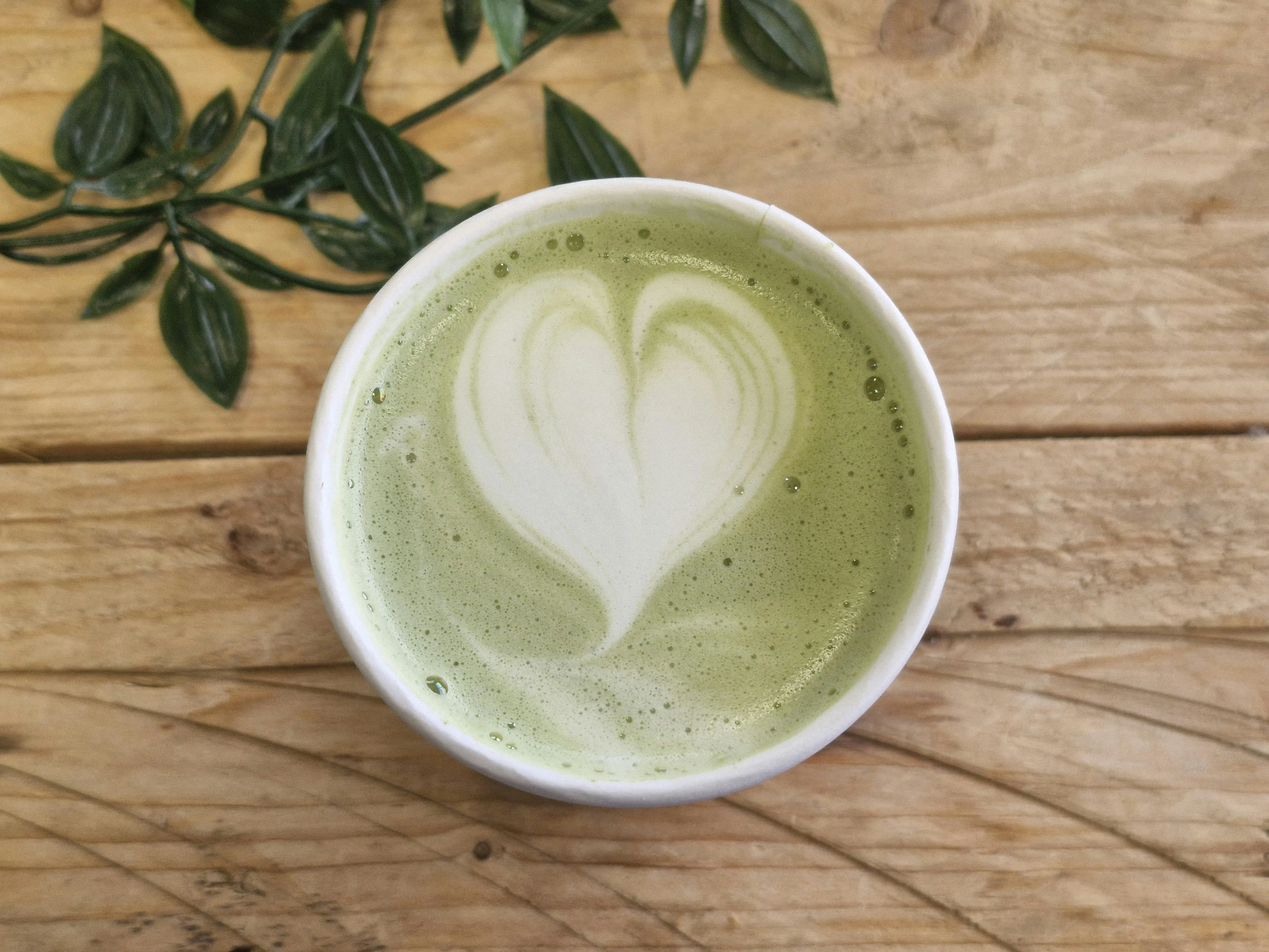 A matcha latte with heart-shaped latte art, served in a white cup on a wooden table with green leaves. Recovery drink.
