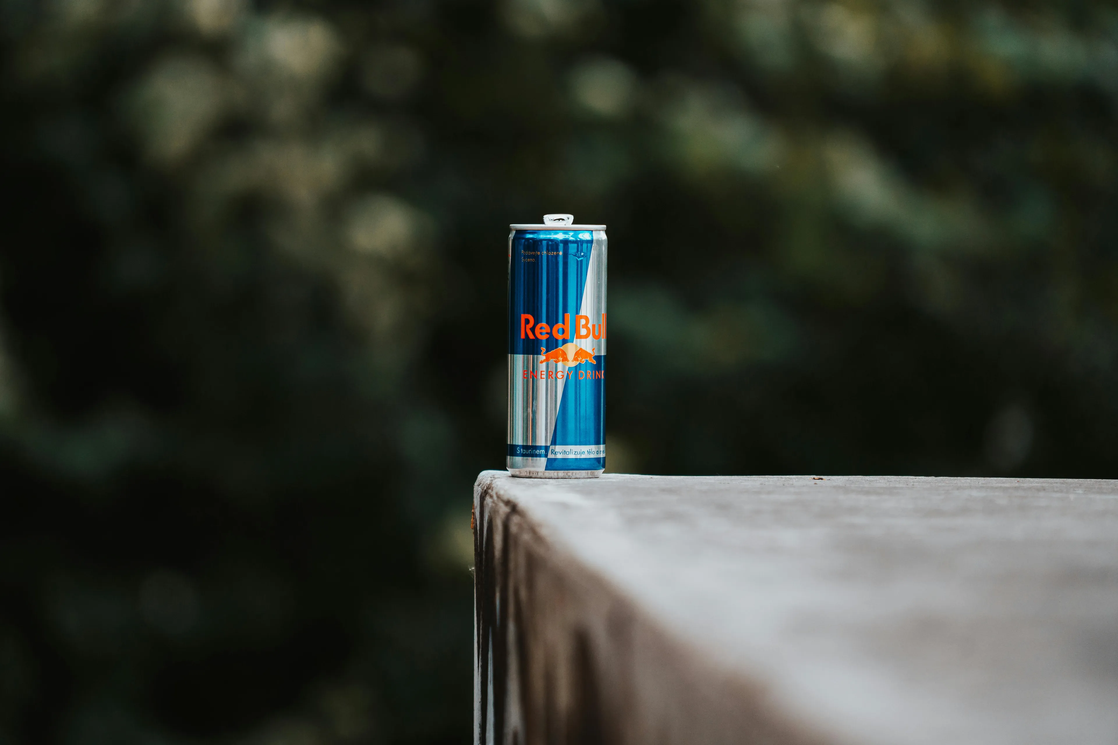 A Red Bull energy drink can stands on a concrete ledge against a soft-focus green background. Recovery drink.