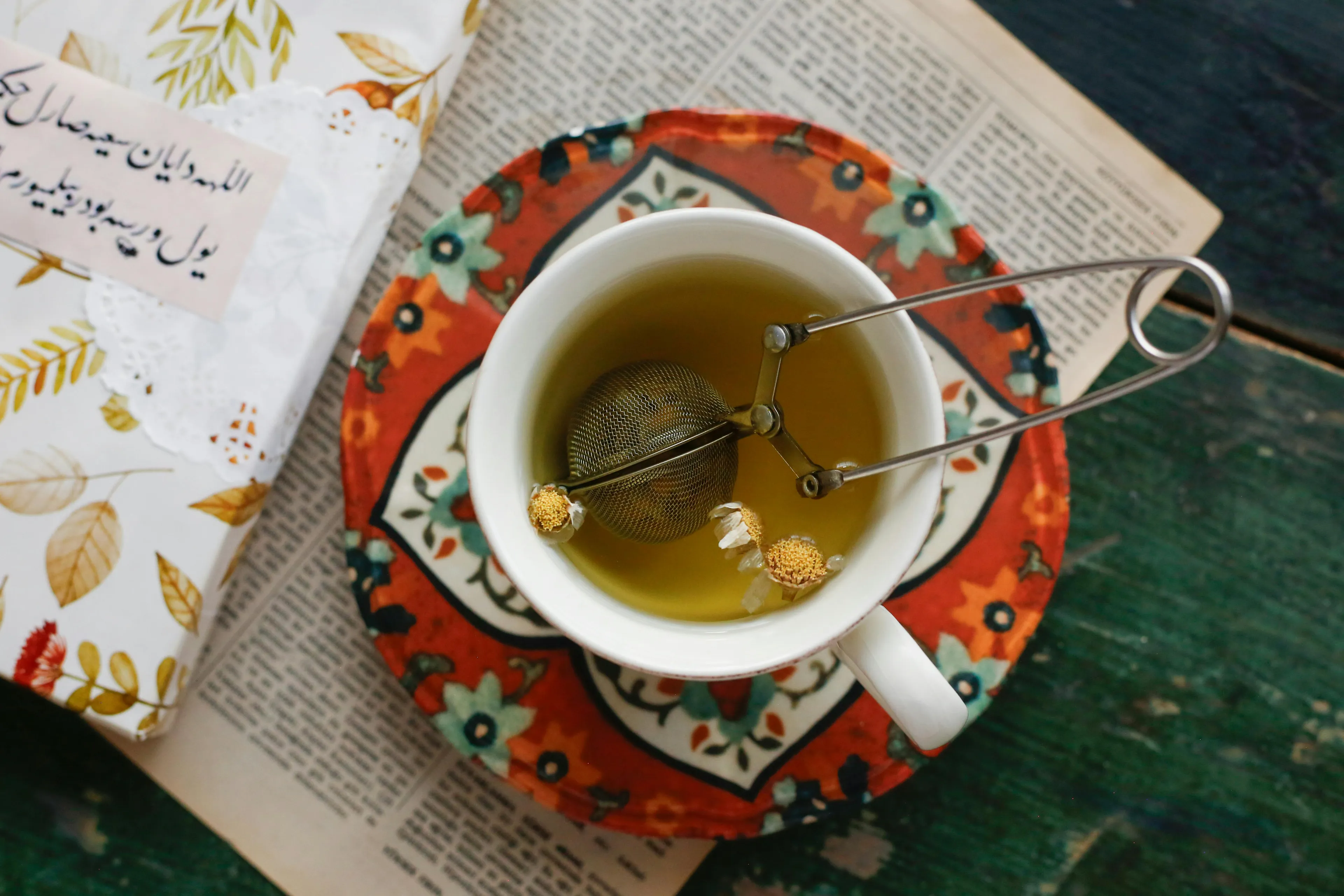 Overhead shot of chamomile tea brewing in a white mug with a tea infuser, on a patterned coaster and newspaper.