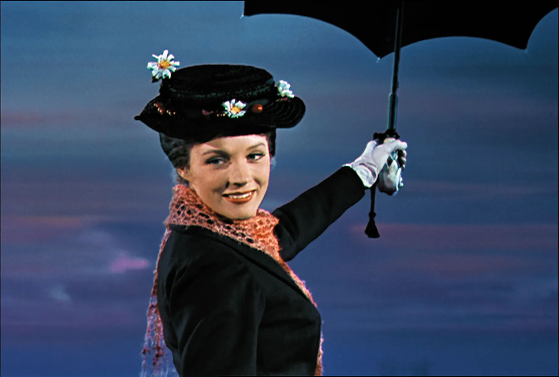 Mary Poppins smiles while holding an open black umbrella against a blue and purple sky, wearing a floral hat.