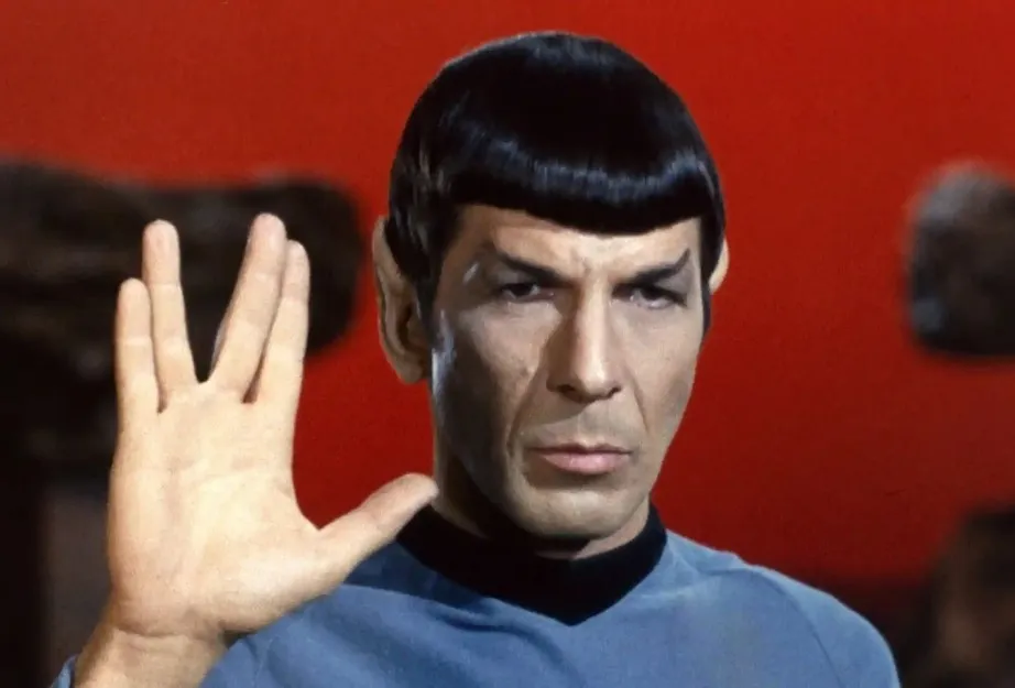 Spock, a fictional character with pointed ears and dark hair, wearing a blue Starfleet uniform and performing the Vulcan salute.