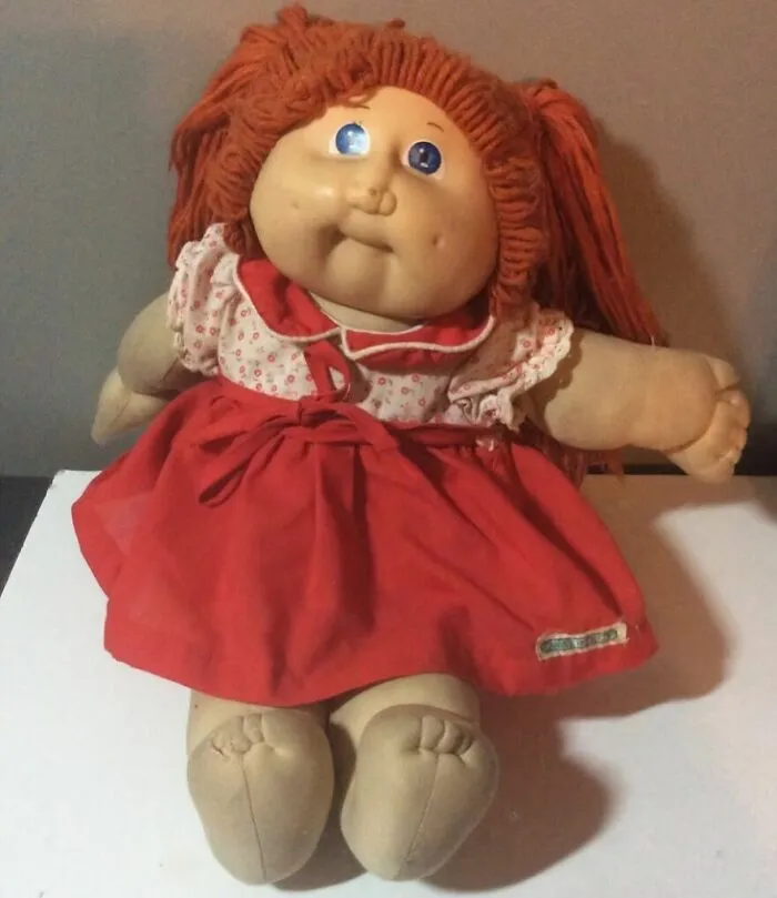 A Cabbage Patch Kid doll with red yarn hair, wearing a white floral shirt and red dress, a trip down memory lane for many.