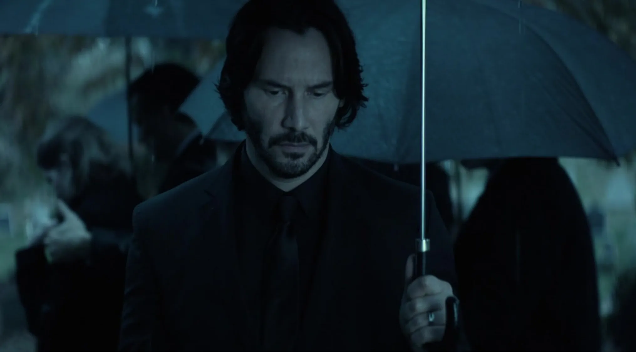 Keanu Reeves, with a beard and wearing a black suit, holding an umbrella in a somber scene. General Knowledge quiz.