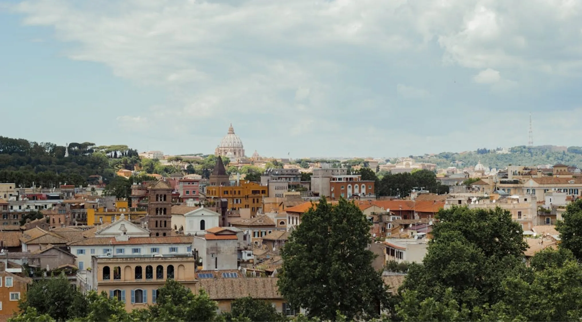 An aerial view of the Rome cityscape under a cloudy sky, showing buildings, trees, and the dome of St. Peter's Basilica. General Knowledge.
