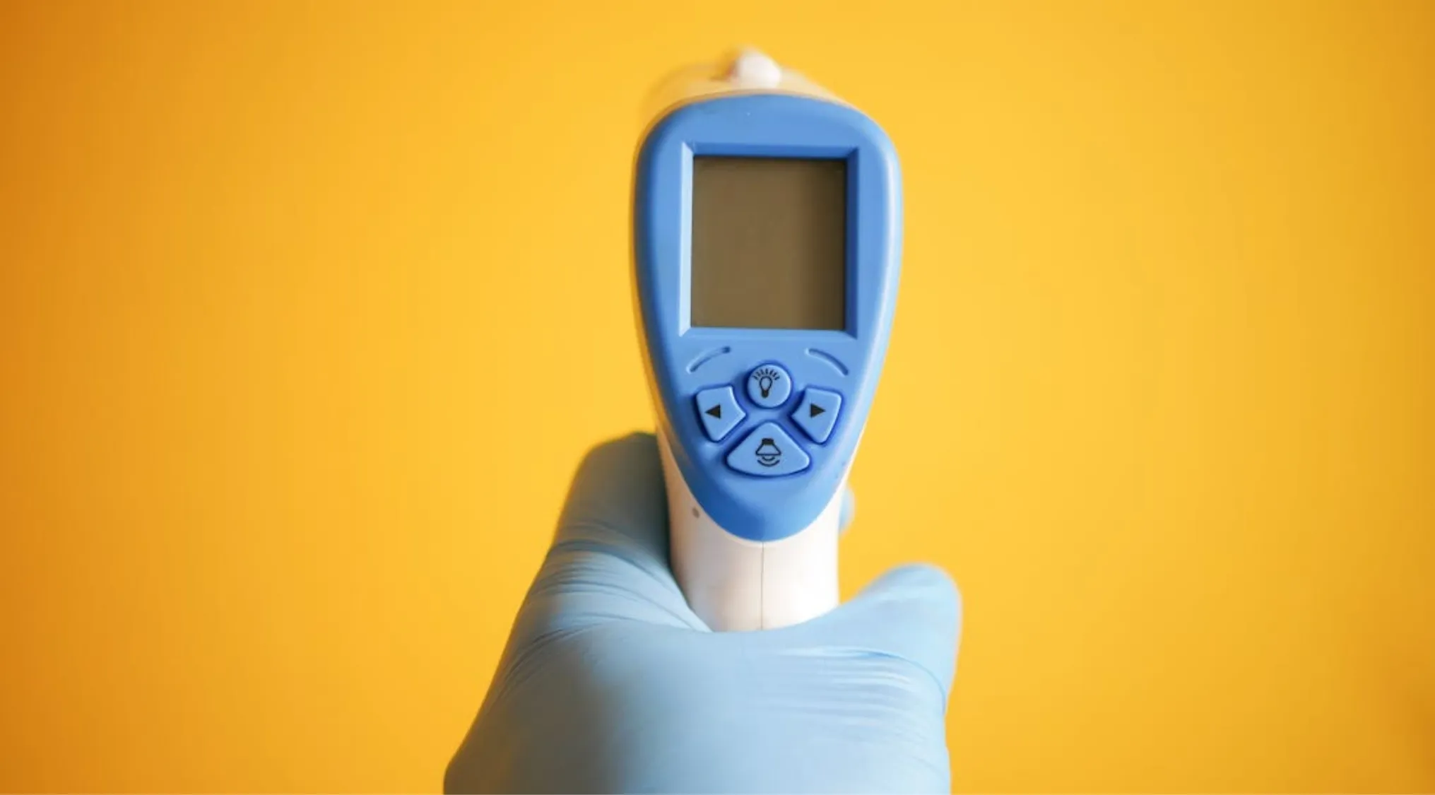 A person in a blue glove holding a blue and white infrared thermometer against a yellow background, for general knowledge.