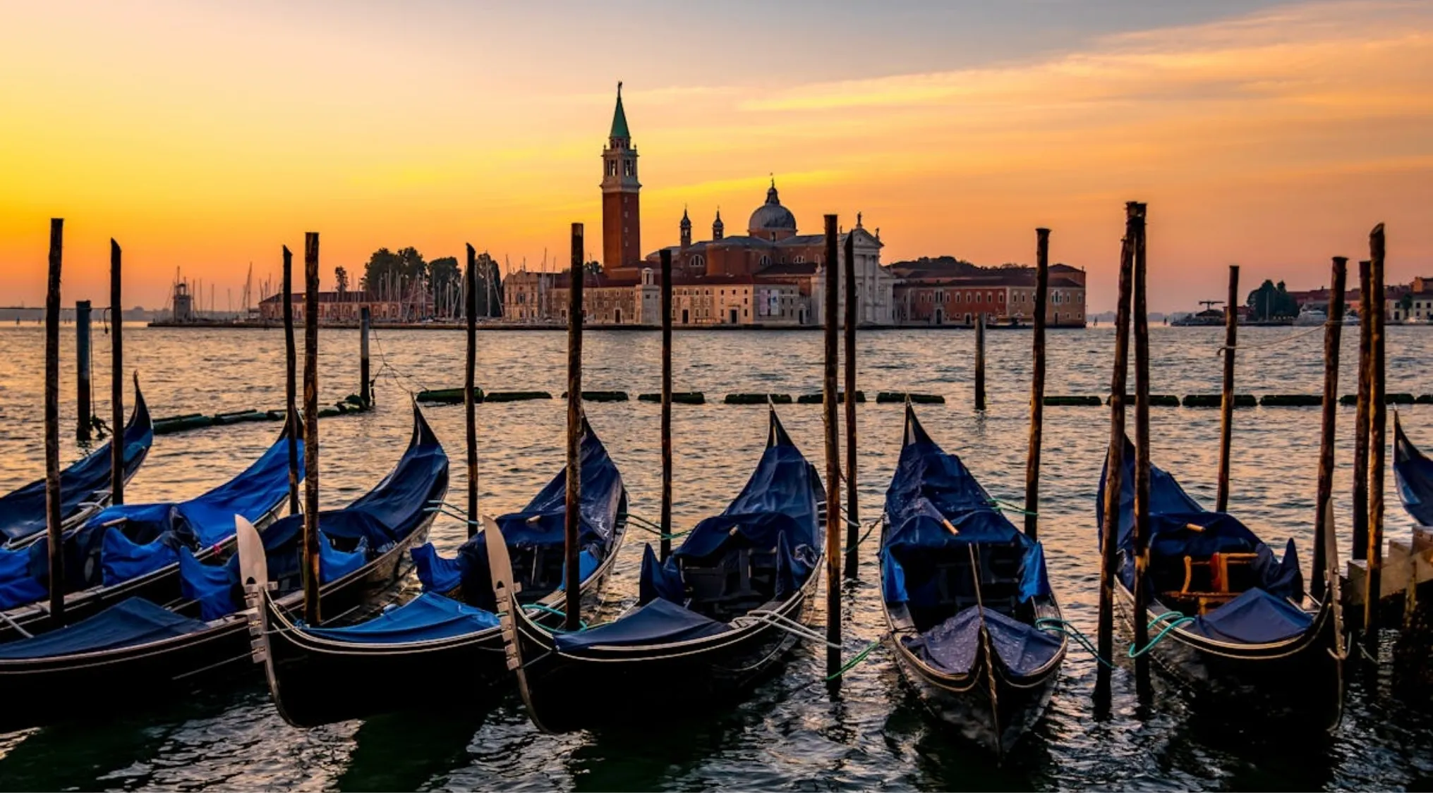 Gondolas moored on calm water at sunset in Venice, Italy, with iconic canals and historic buildings in the background. A general knowledge question answered by this famous Italian city.