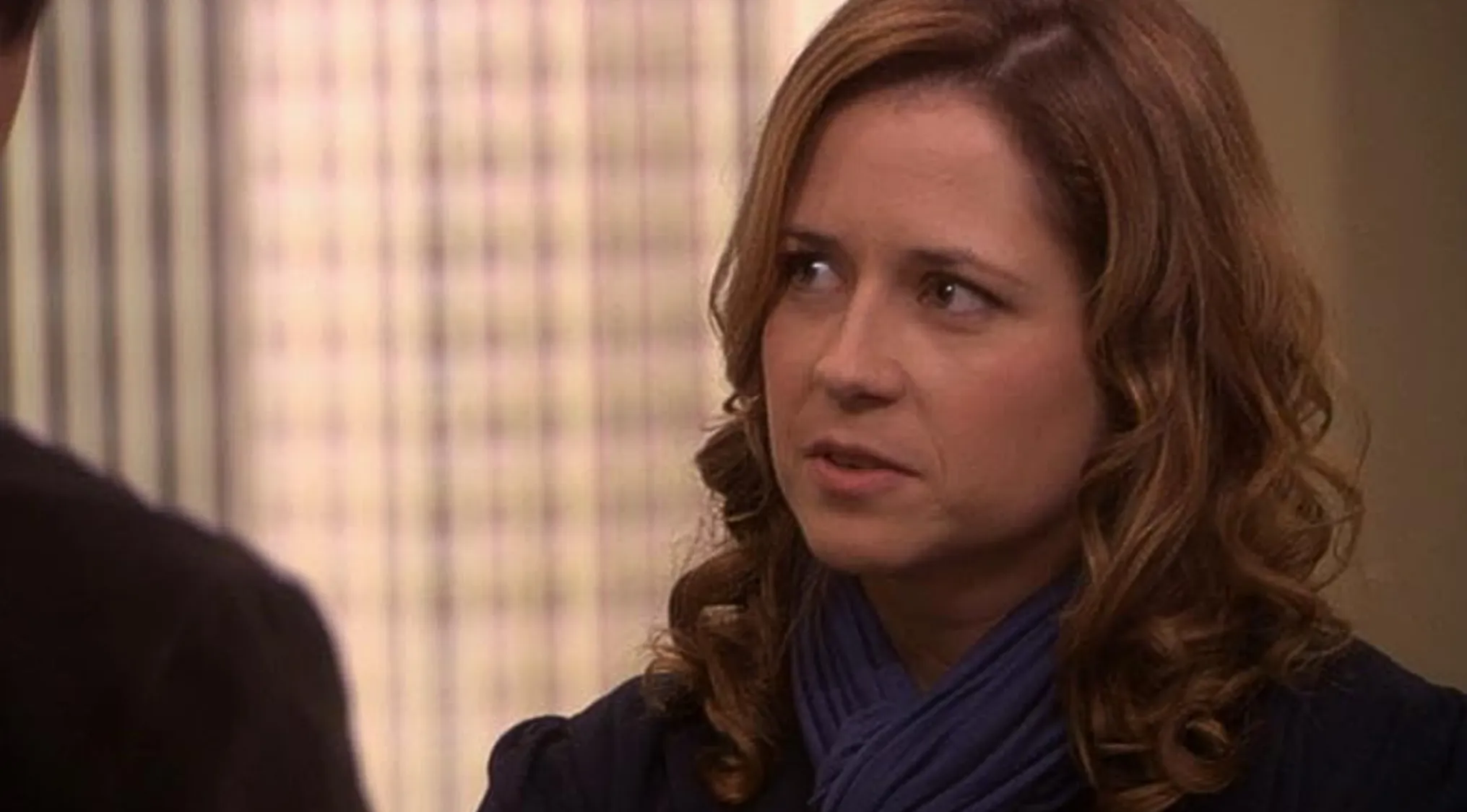 Pam Beesly from The Office Quiz, looking away with a thoughtful expression. She wears a blue scarf and dark top.