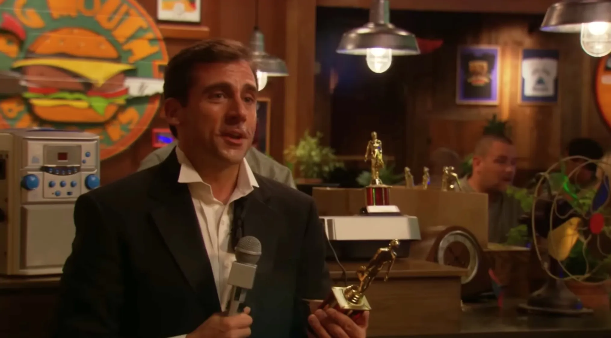 Michael Scott hosting The Office Quiz awards show, holding a microphone and a "Dundie" award.