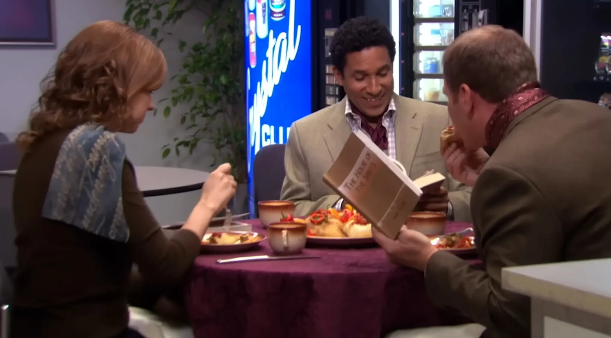 The Office Quiz: Toby, Pam, and Oscar at a small table, eating and Oscar reading from a book, forming a club.
