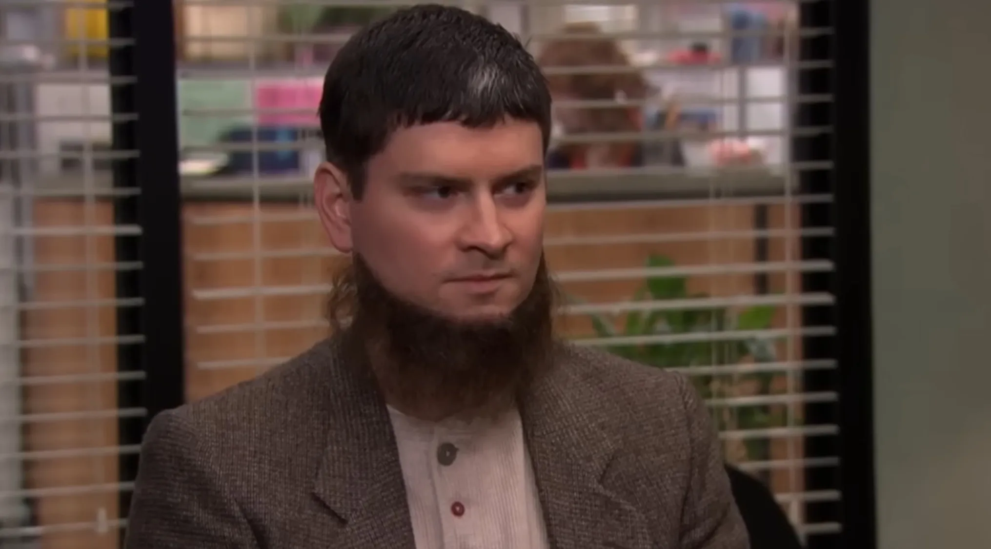 Dwight's cousin, Mose, from The Office Quiz, with a distinctive haircut and beard, sitting in an office setting.