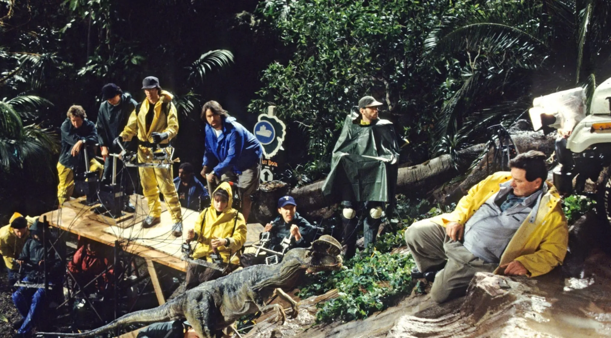 BTS photo showing the crew and a raptor animatronic on a rainy set. Guess the movie franchise!