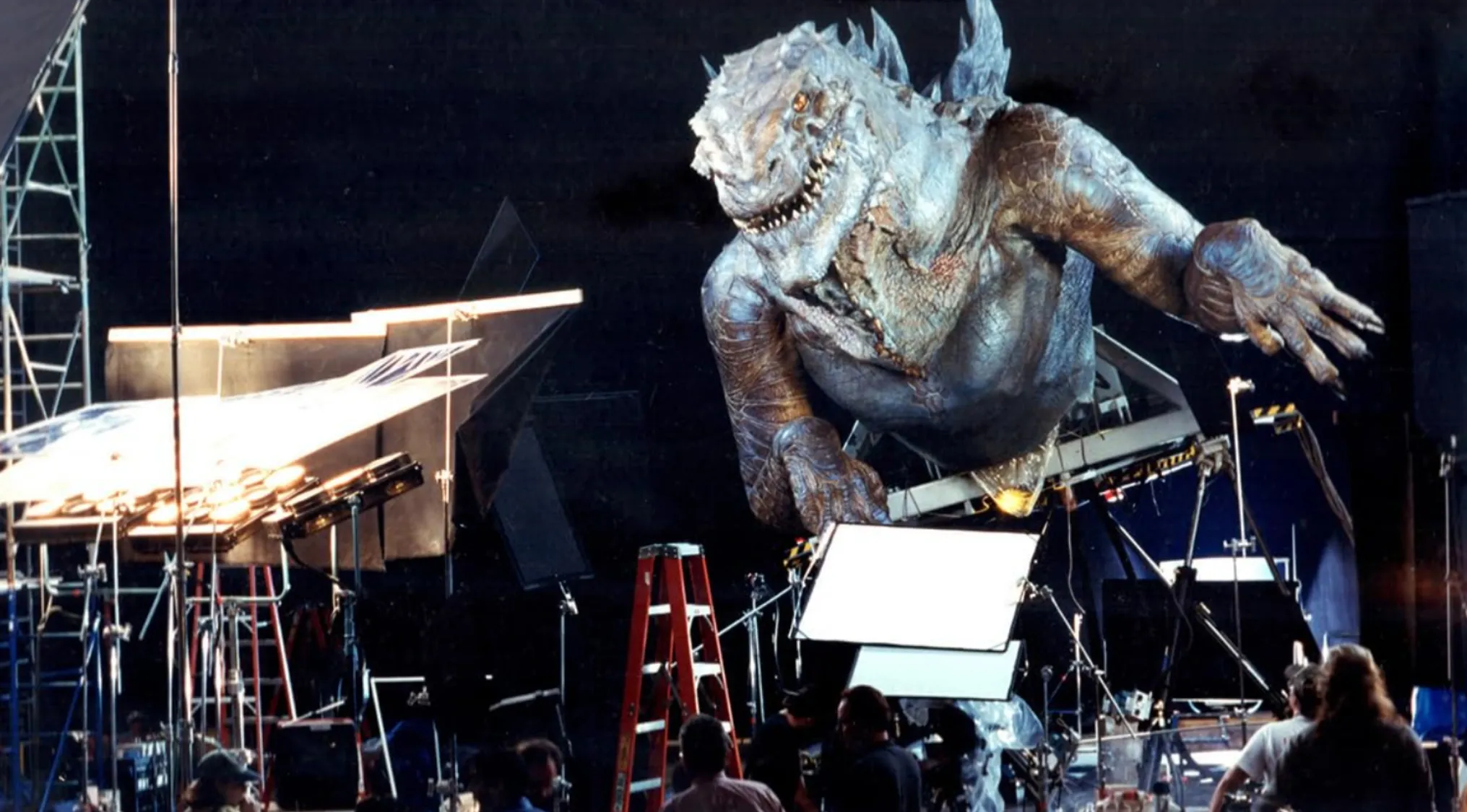 BTS image of a large lizard-like film creature on a dark set with production crew, lights, and equipment. Guess the movie.