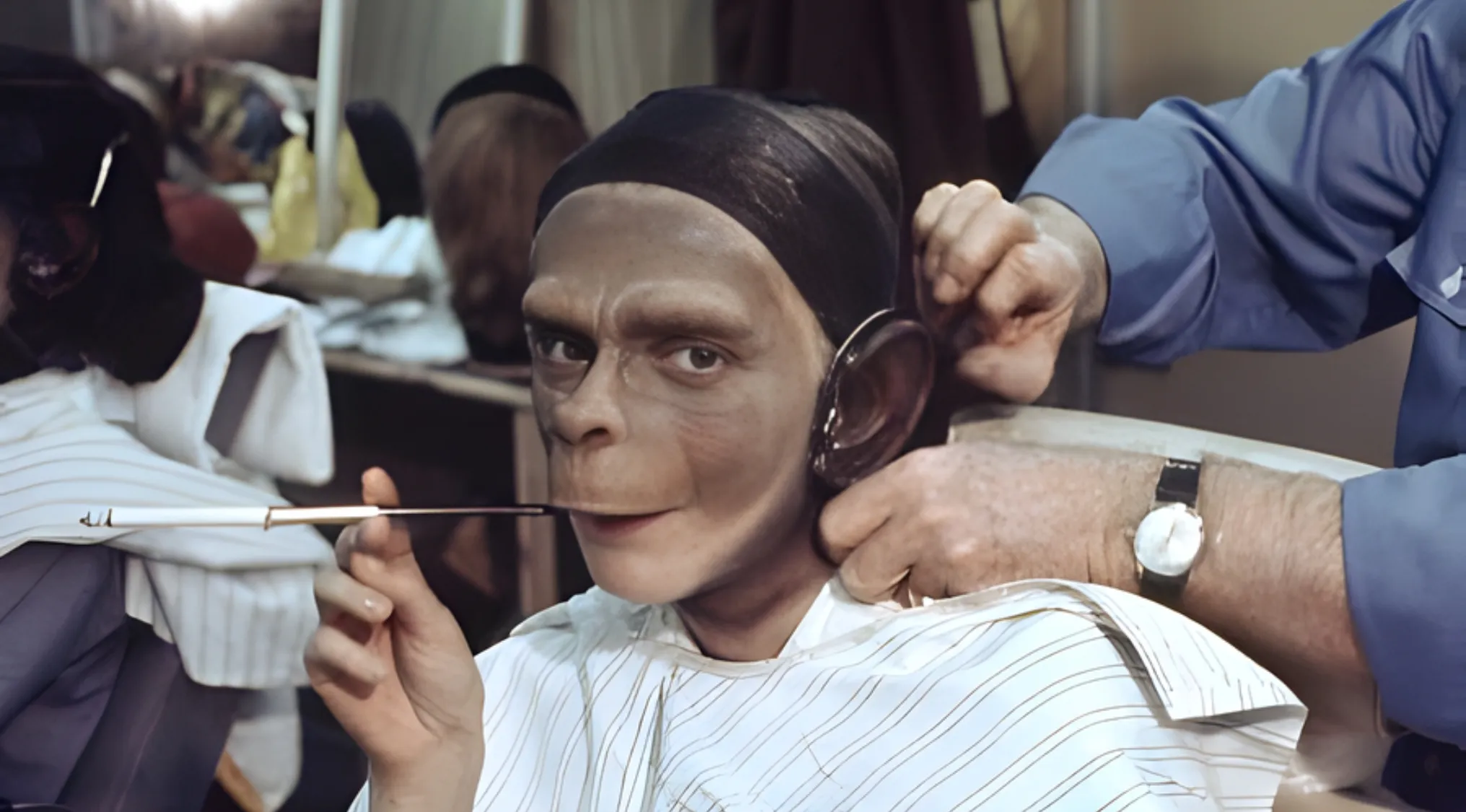 Guess The Movie from this BTS image. An actor in ape makeup is having an ear adjusted, holding a cigarette holder.