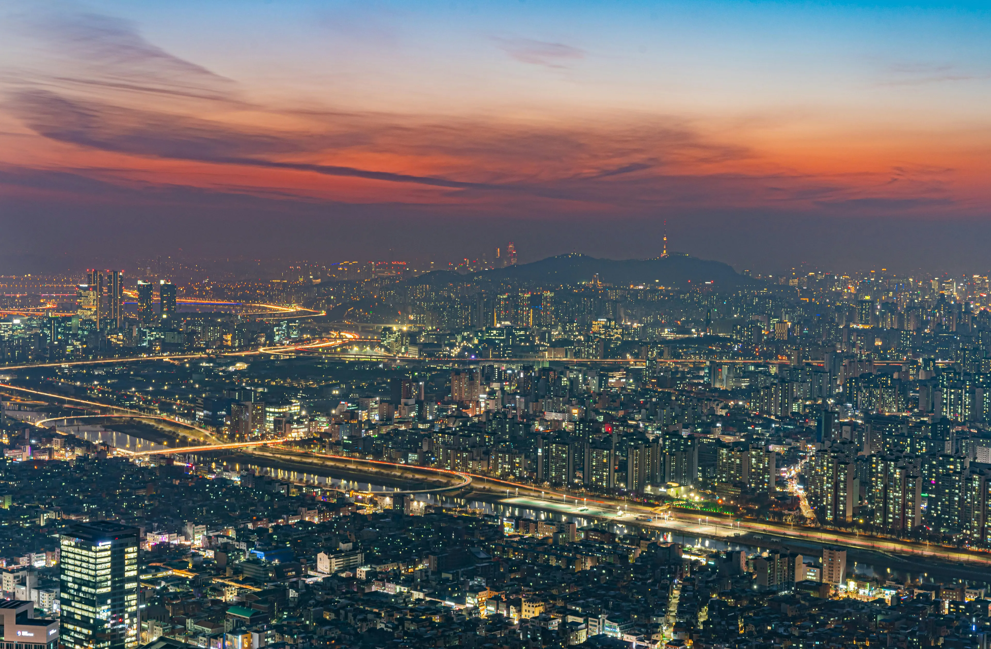 General Knowledge: Elevated night view of the capital of South Korea, Seoul, with illuminated buildings and roads under a colorful sunset.