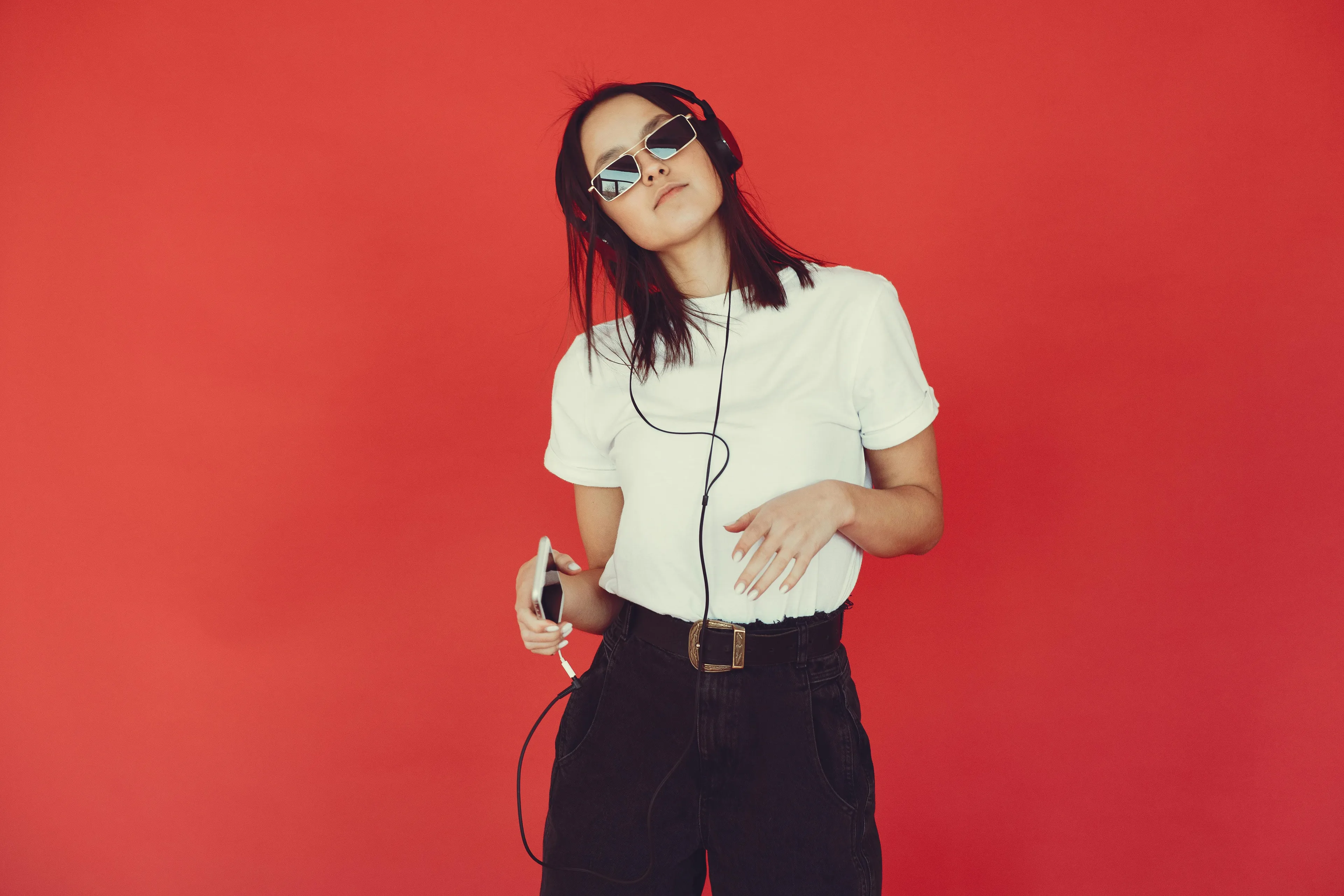 A young woman with sunglasses and headphones, holding a phone, on a red background. Explore general knowledge in pop music.