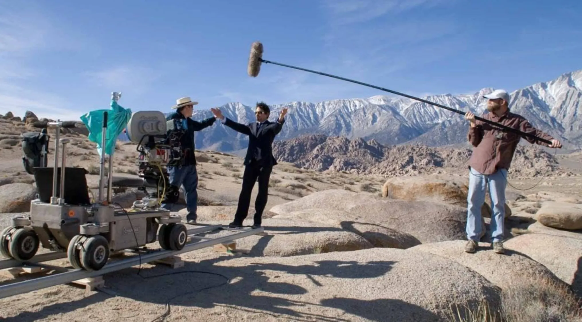 BTS image of a Movie production in a rocky, mountainous desert. An actor in a suit stands on camera tracks.