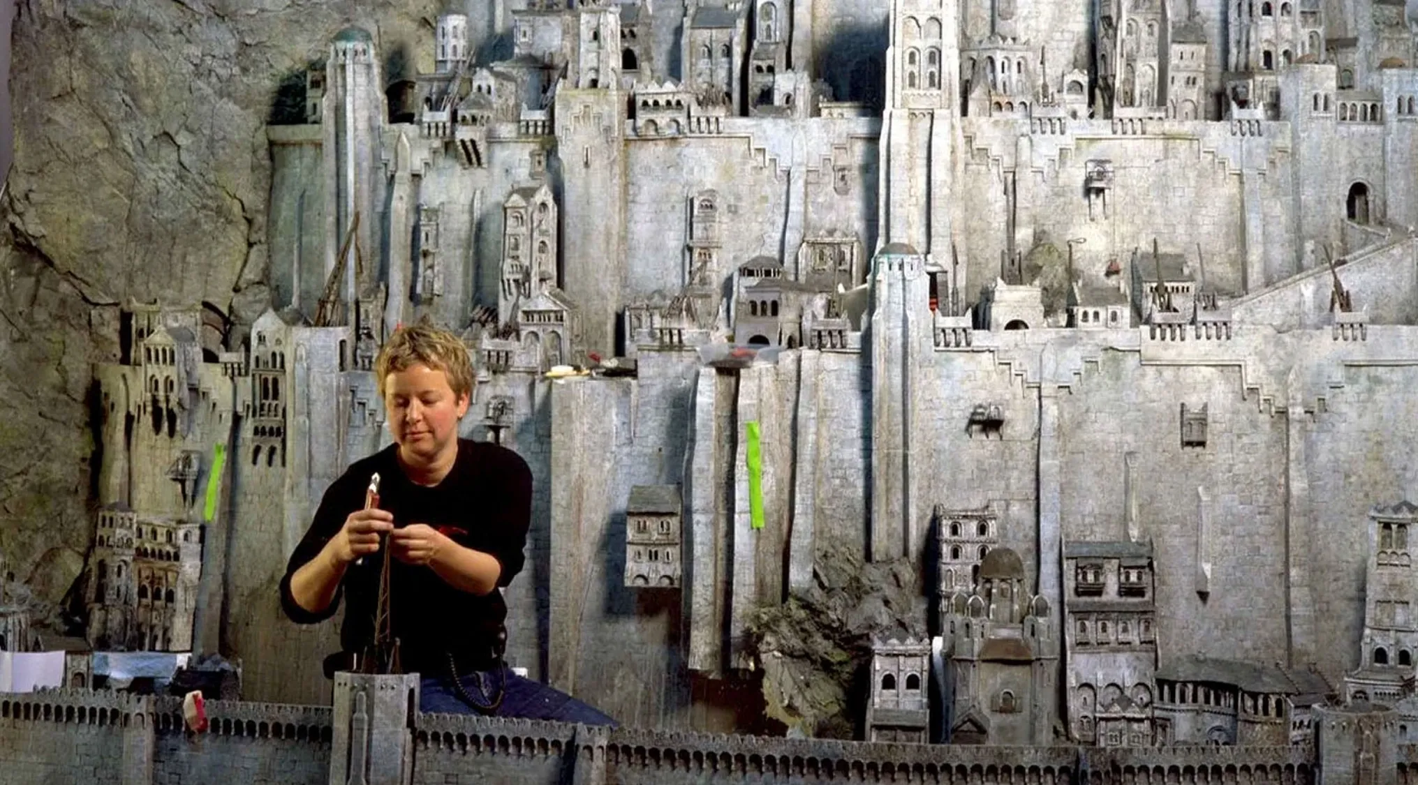 A person works on a large, detailed miniature movie set, constructing parts of a massive stone city. Guess the movie franchise from this BTS.