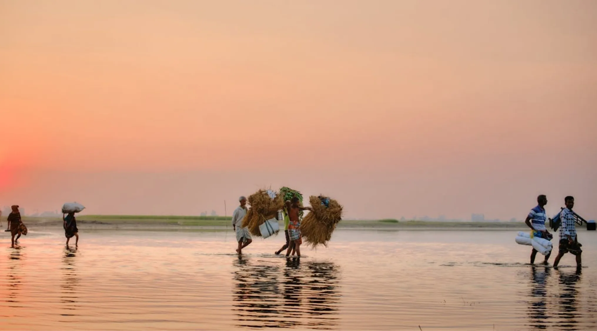 People walking through shallow water at sunset, carrying bundles. This relates to non-European migrants to Australia.