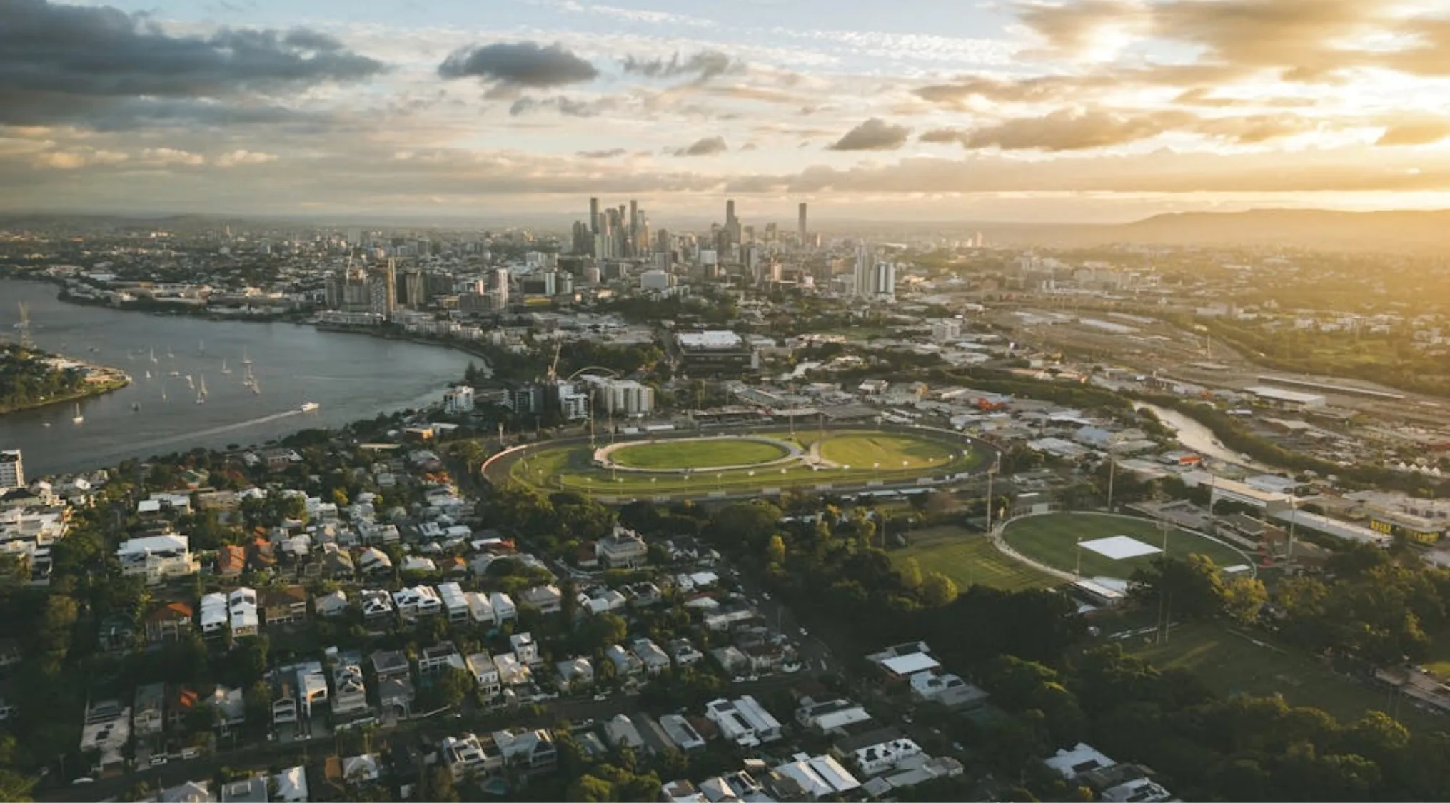 Aerial view of Brisbane at sunset, showing the river, buildings, sports fields, and residential areas. The capital of Queensland.