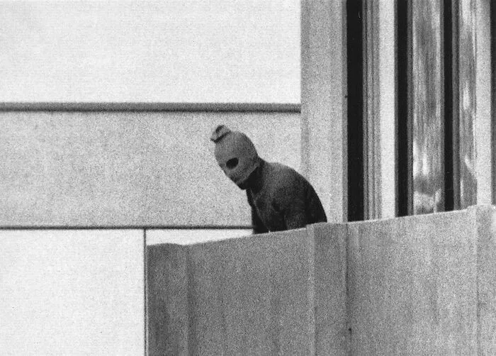 Black and white photo from 1960 to 2010 showing a person in a mask on a building balcony during a notable event.