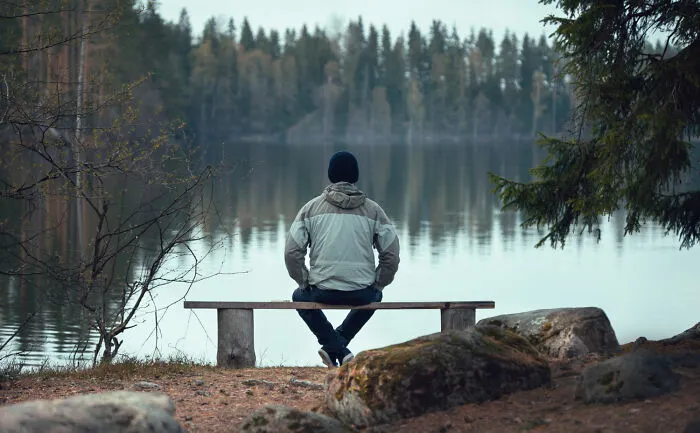 Person sitting alone on a bench by a calm lake, reflecting on moments of being a good person ending in betrayal.