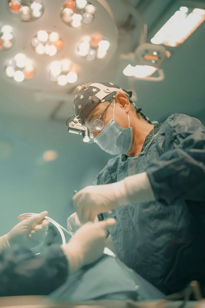 A surgeon in scrubs, mask, and headlamp working under bright operating room lights. Highlights patients being ignored in emergencies.