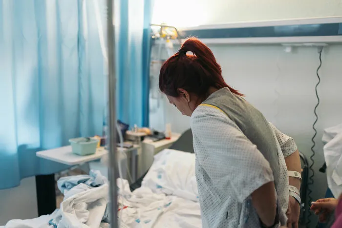 A patient with red hair in a hospital gown, seen from behind, standing next to a bed in a ward, ignored in emergencies.