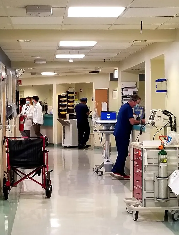 A busy hospital hallway with medical staff, a red wheelchair, and equipment. Highlights patients being ignored in emergencies.