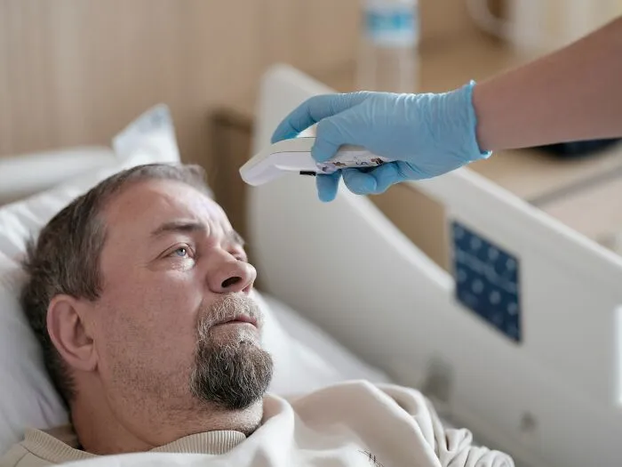 A patient in a hospital bed having his temperature taken by a gloved hand. Stories of patients being ignored in emergencies.