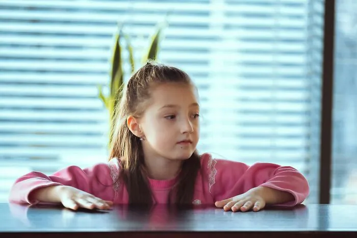 Young girl in a pink sweater displaying intelligence and curiosity while sitting at a table indoors with blinds in the background.